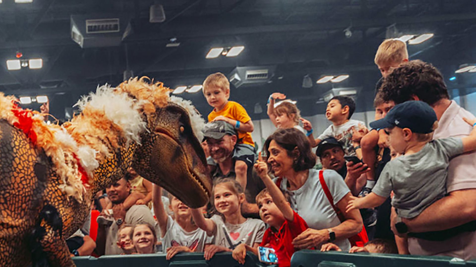 Children and adults excitedly reaching out to touch a life-like dinosaur puppet with colorful feathers on its head behind a barrier in an indoor event.