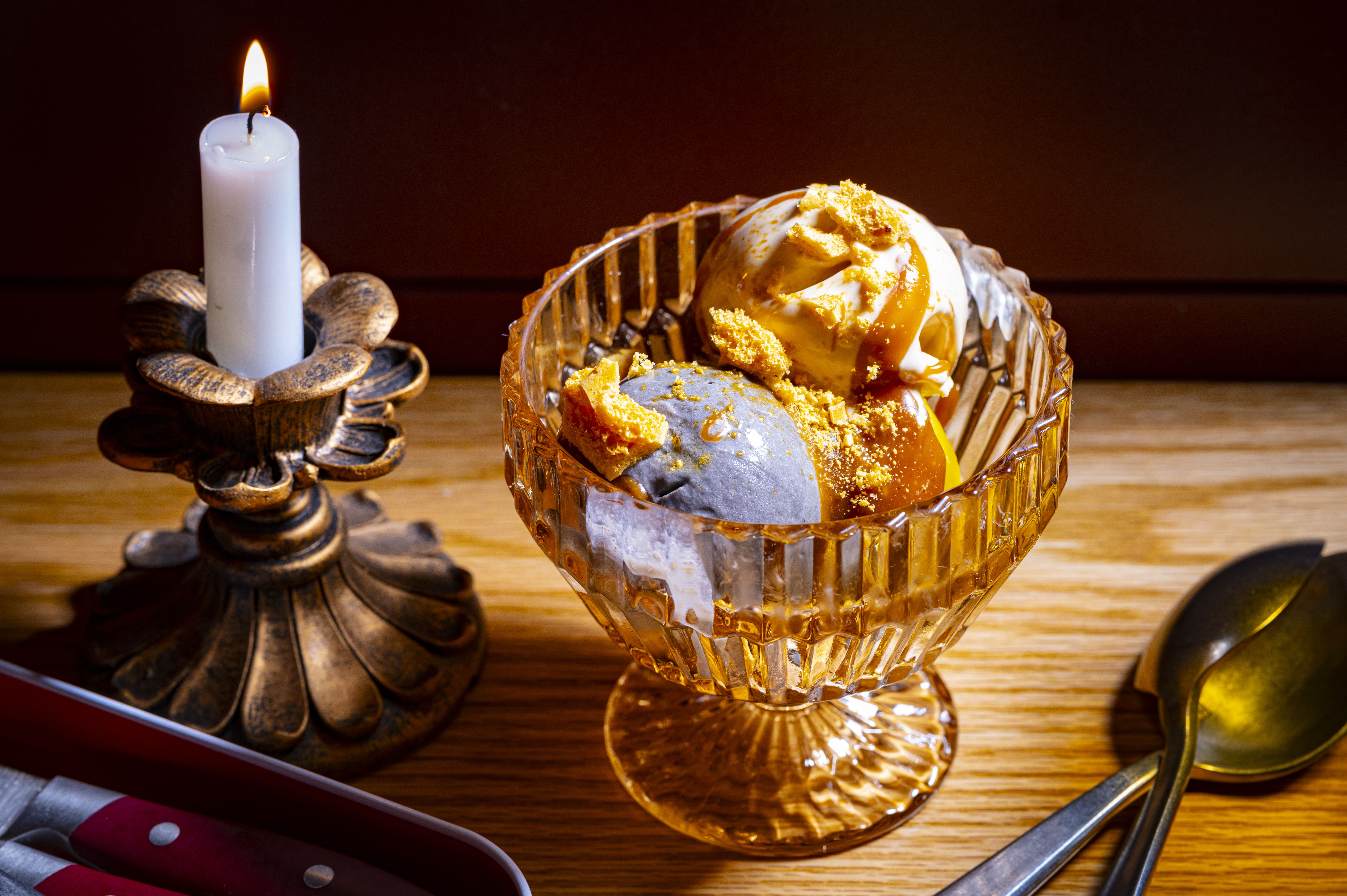 A lit white candle in an ornate metal holder sits beside a crystal dessert bowl filled with caramel-drizzled ice cream scoops and toffee crumbs on a wooden table.