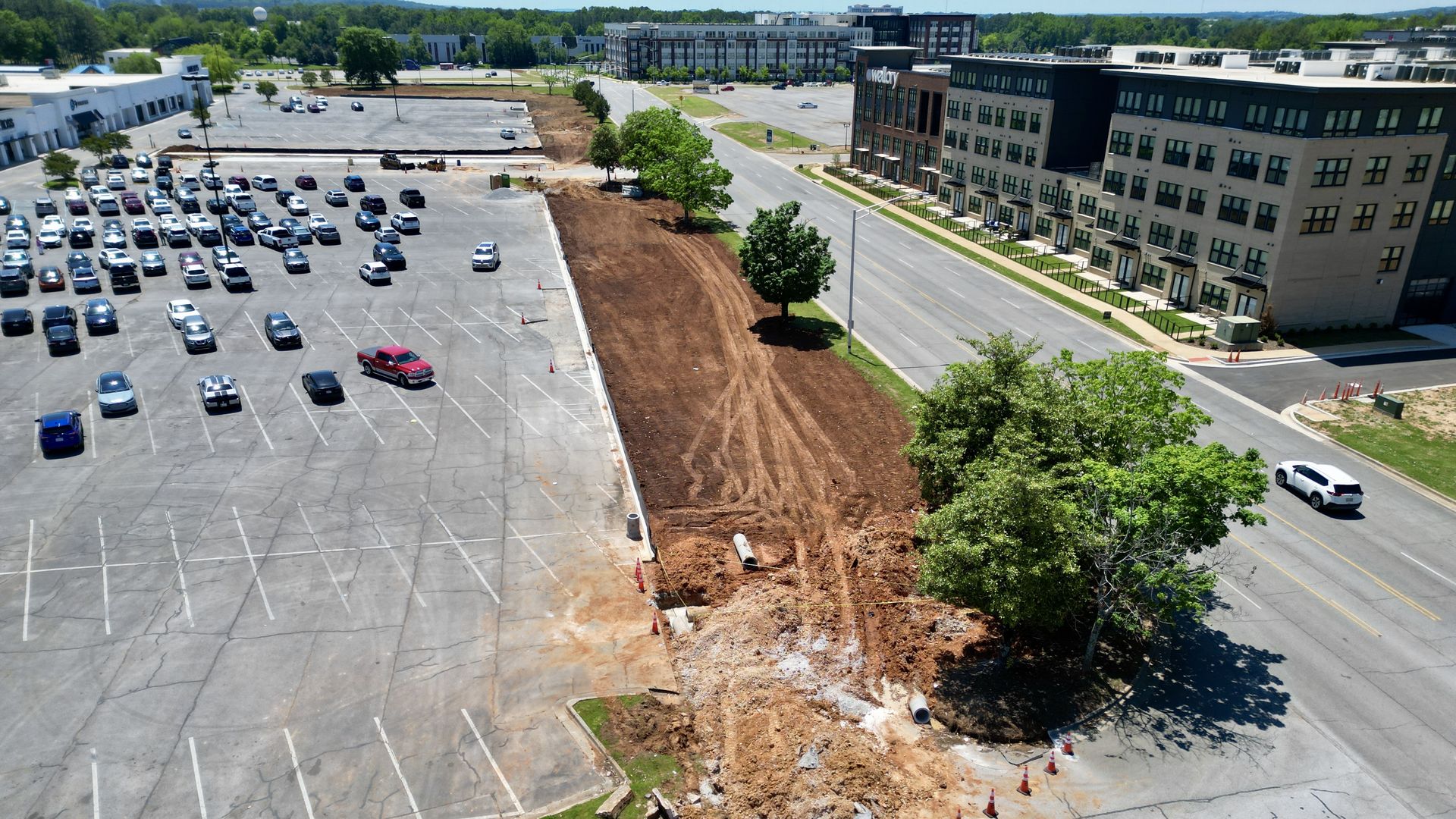 An aerial view of a large parking lot filled with cars beside a dirt construction zone with orange barriers, and a modern multi-story office building to the right; trees and hills in the distance.