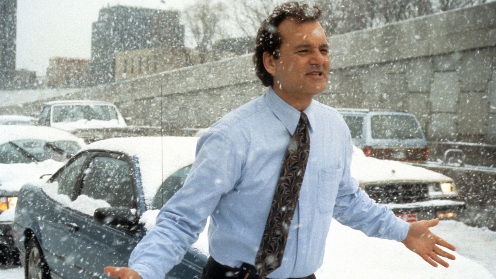 Bill Murray in a scene from "Groundhog Day," wearing light blue shirt and patterned tie standing with arms open in heavy snowfall on a snow-covered street with cars and buildings in the background.
