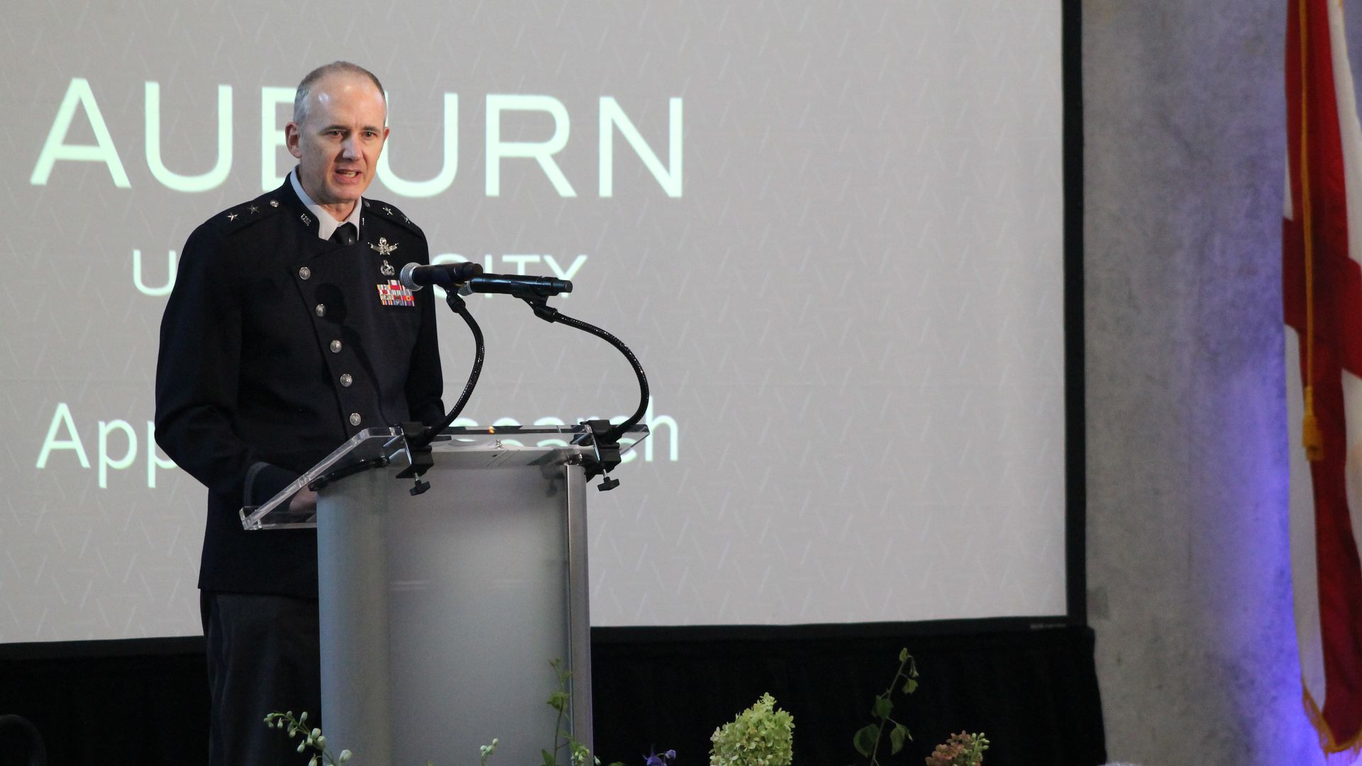 Senior military officer in a dark dress uniform speaks at a podium with two microphones; a background screen reads "AUBURN UNIVERSITY." Colorful flowers line the stage, with flags on the right.