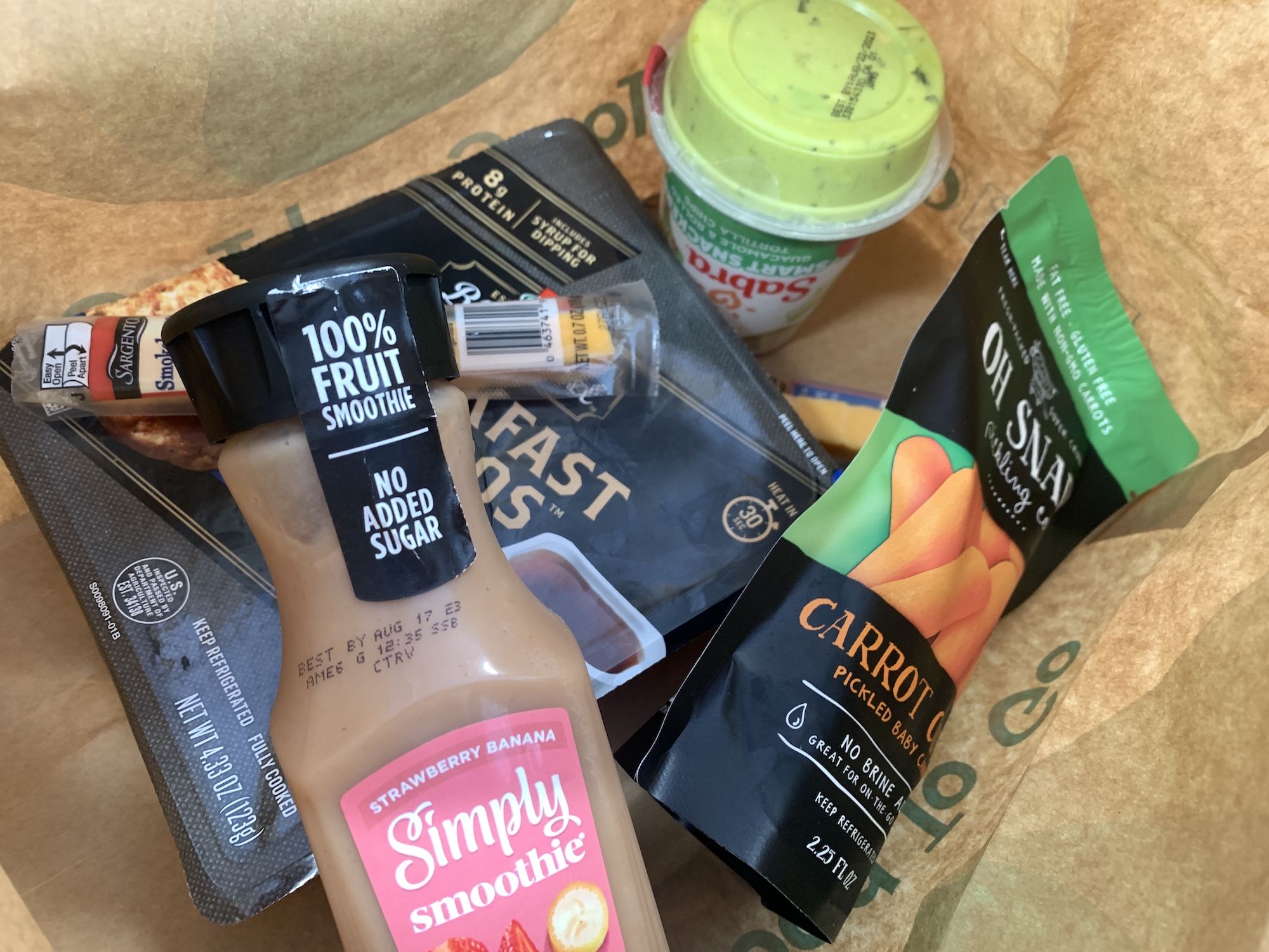 Smoothie, bag of carrots, cheese stick and other snacks in a brown paper bag.