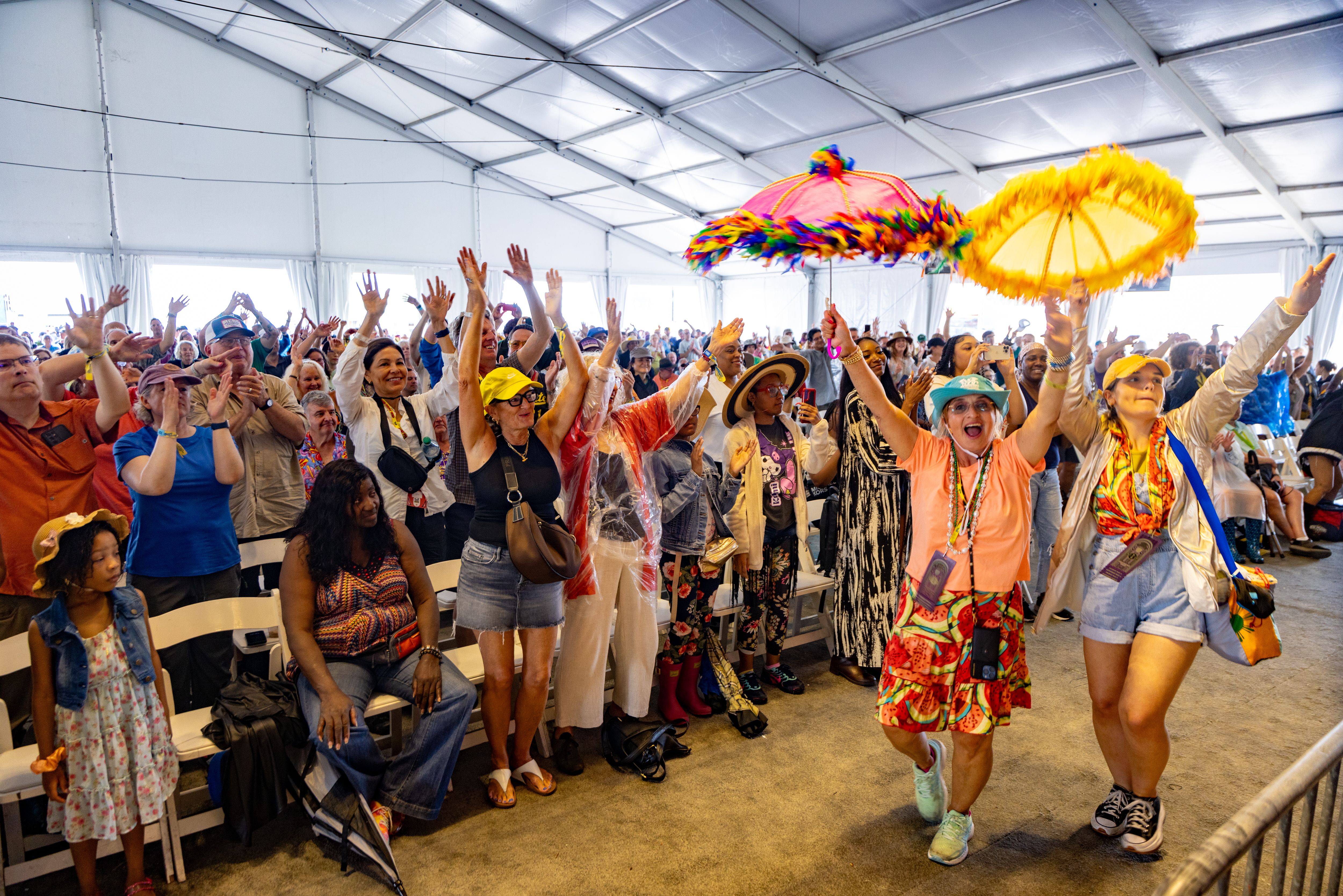 Two women with second-line umbrellas dance at the front of an audience, who's all on their feet with hands in the air.