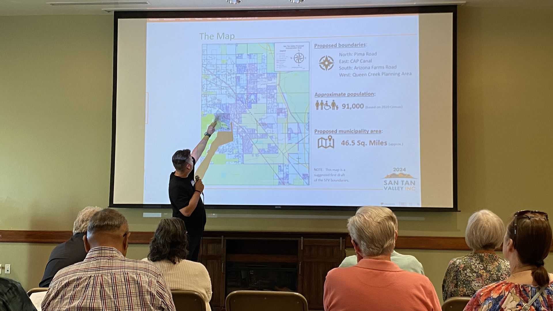 A man points to a map on a large screen while people in folding chairs watch.