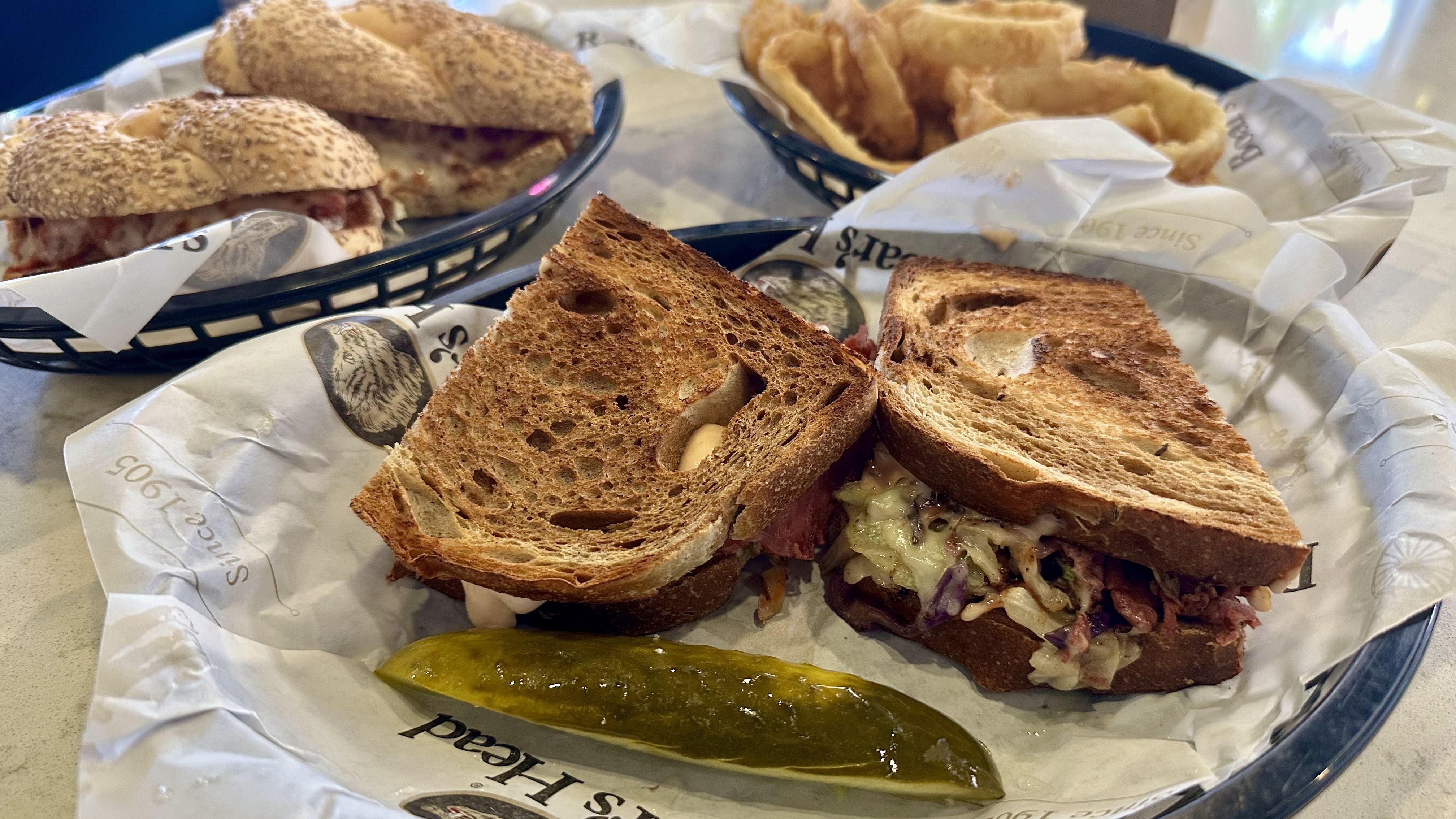 Toasted sandwich with coleslaw and meat on rye bread, served with a pickle spear on branded paper. In the background, a chicken parm sandwich and a basket of onion rings.