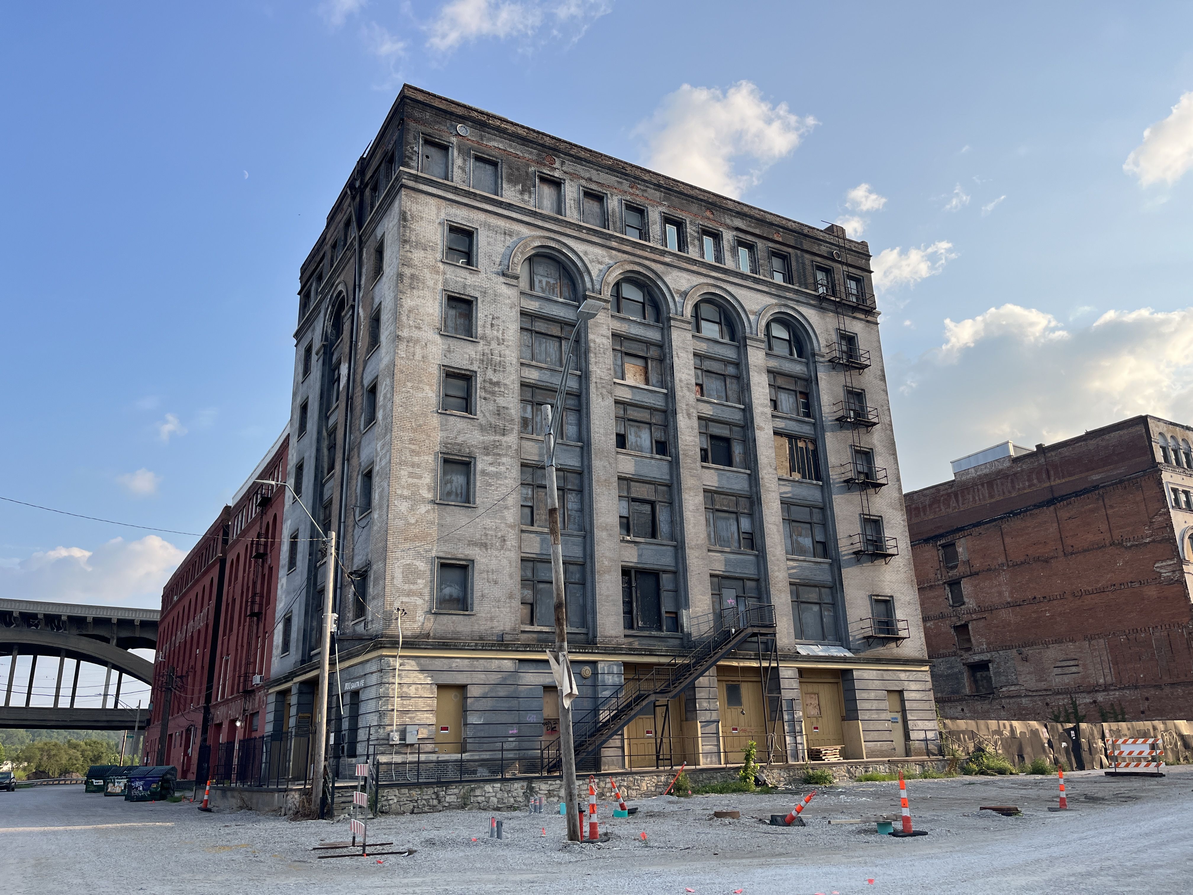 Large old industrial building with boarded windows and fire escape stairs, surrounded by gravel lot and orange construction cones, under a blue sky with scattered clouds.
