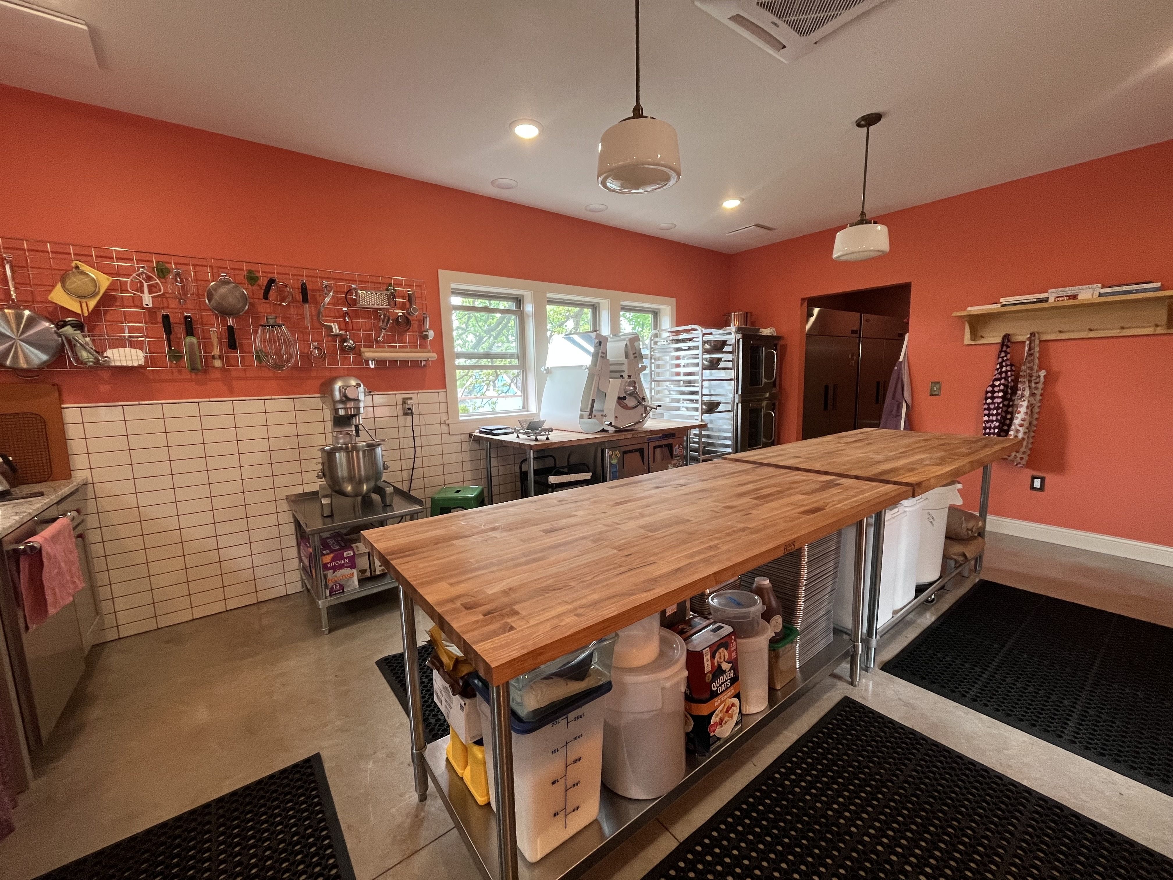 Bright orange kitchen studio with two wooden prep tables, a wall grid of hanging pots and utensils, stainless appliances, and a back area with a fridge; large windows let in light.
