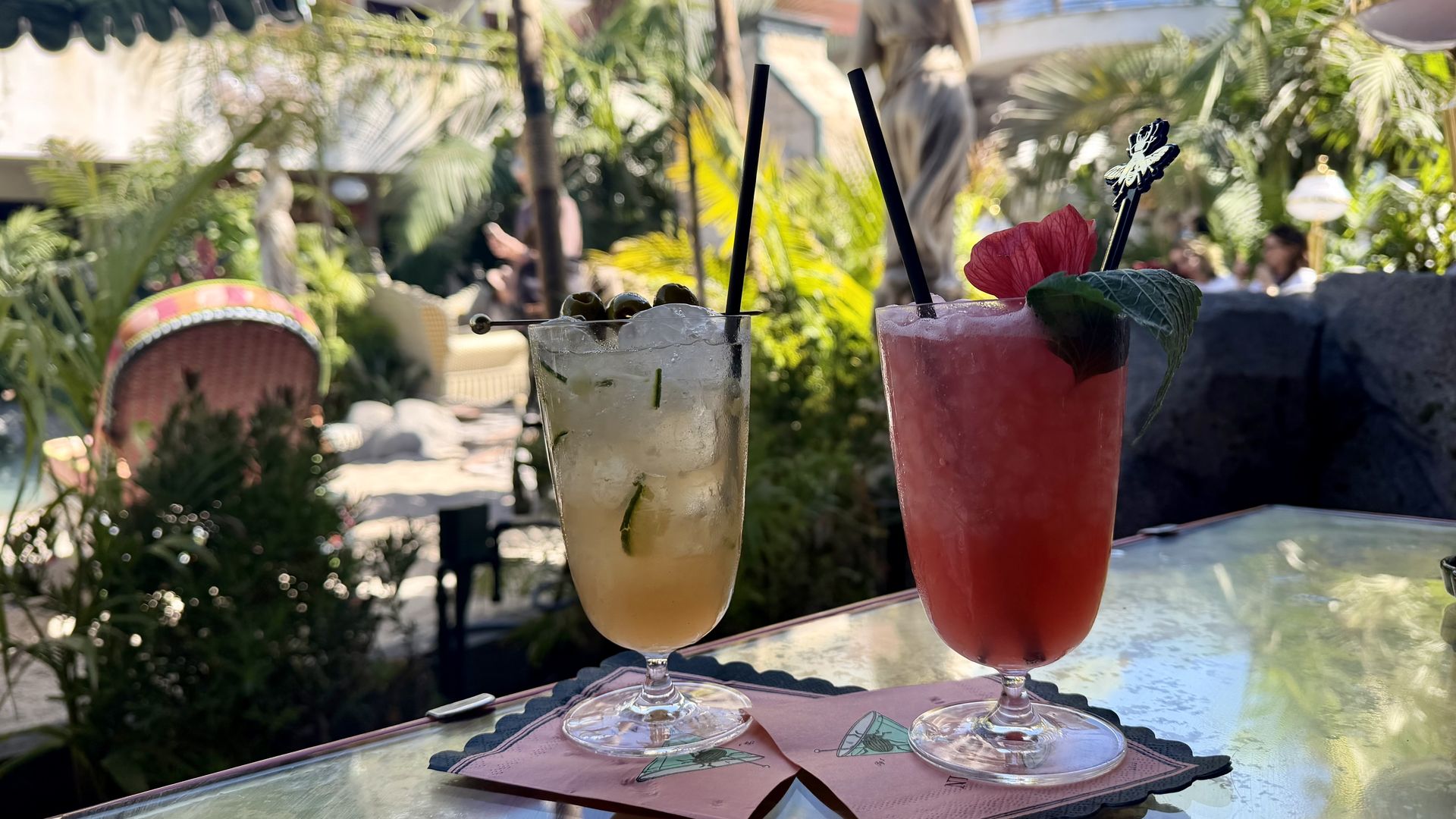 Two tropical cocktails on a glass table outdoors: left pale yellow drink with mint, right pink-red drink with a hibiscus flower and leaf; black straws, pink napkins, lush garden backdrop.