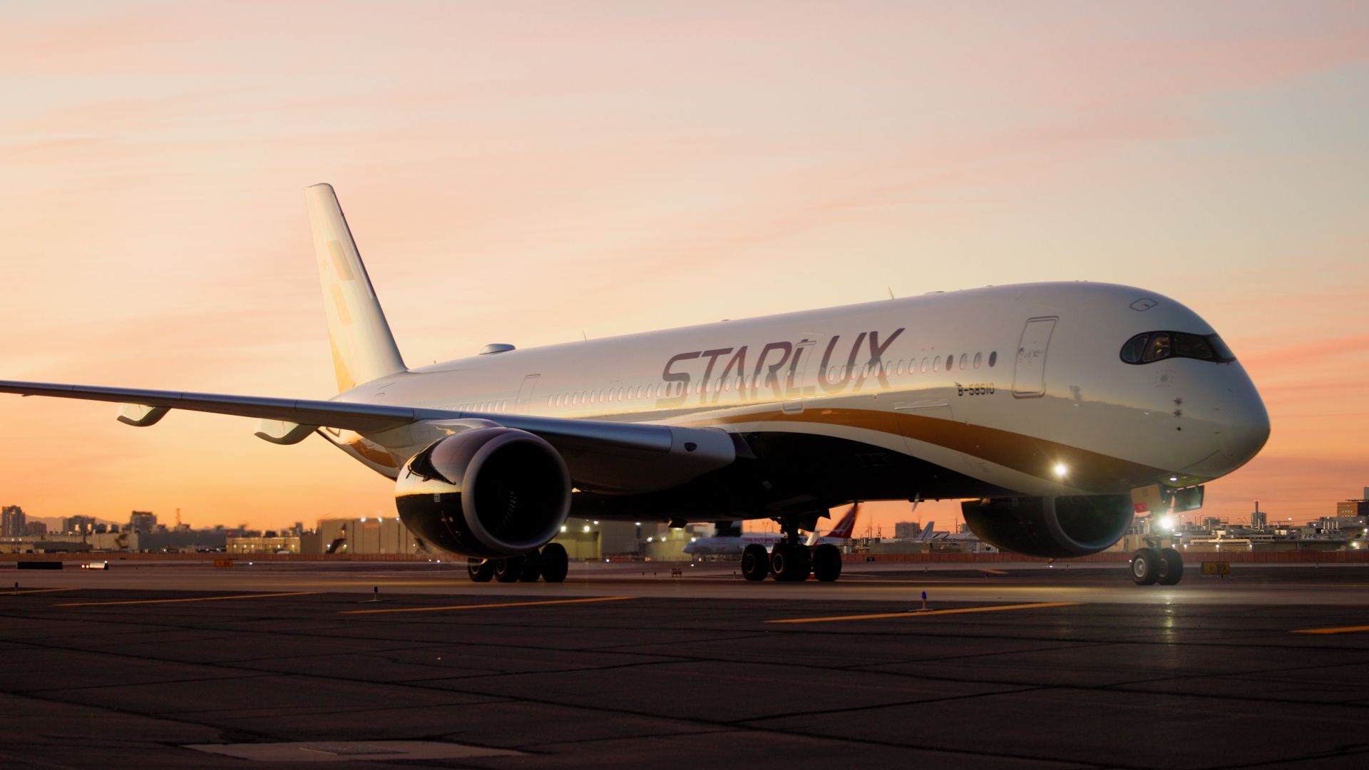 White and gold STARLUX commercial airplane on runway at sunset with city skyline in the background and light reflecting off the engine and nose.
