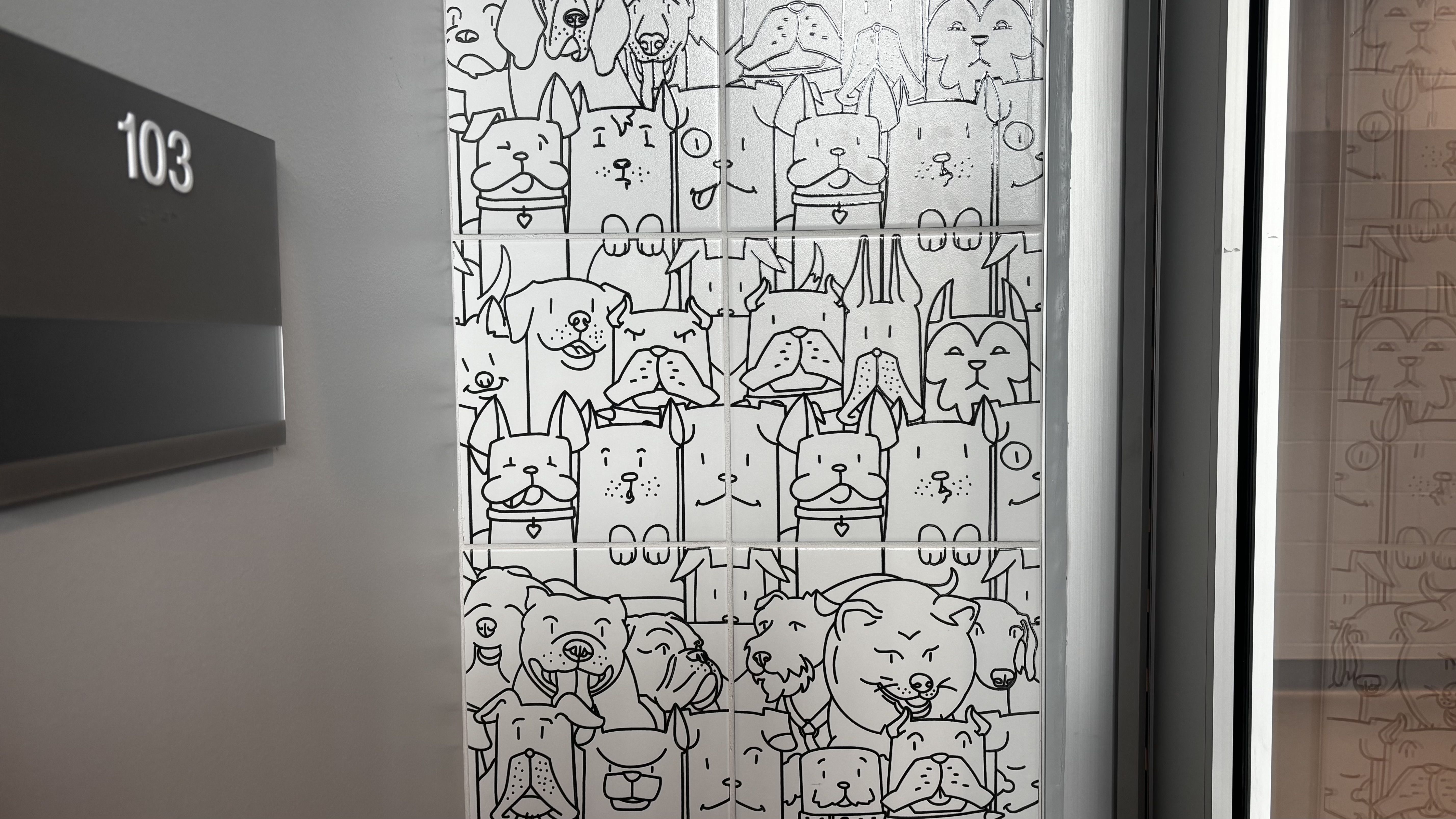 Elevator interior wall covered with a grid of black-and-white cartoon dog faces on tiled panels; a dark panel on the left shows the number 103.