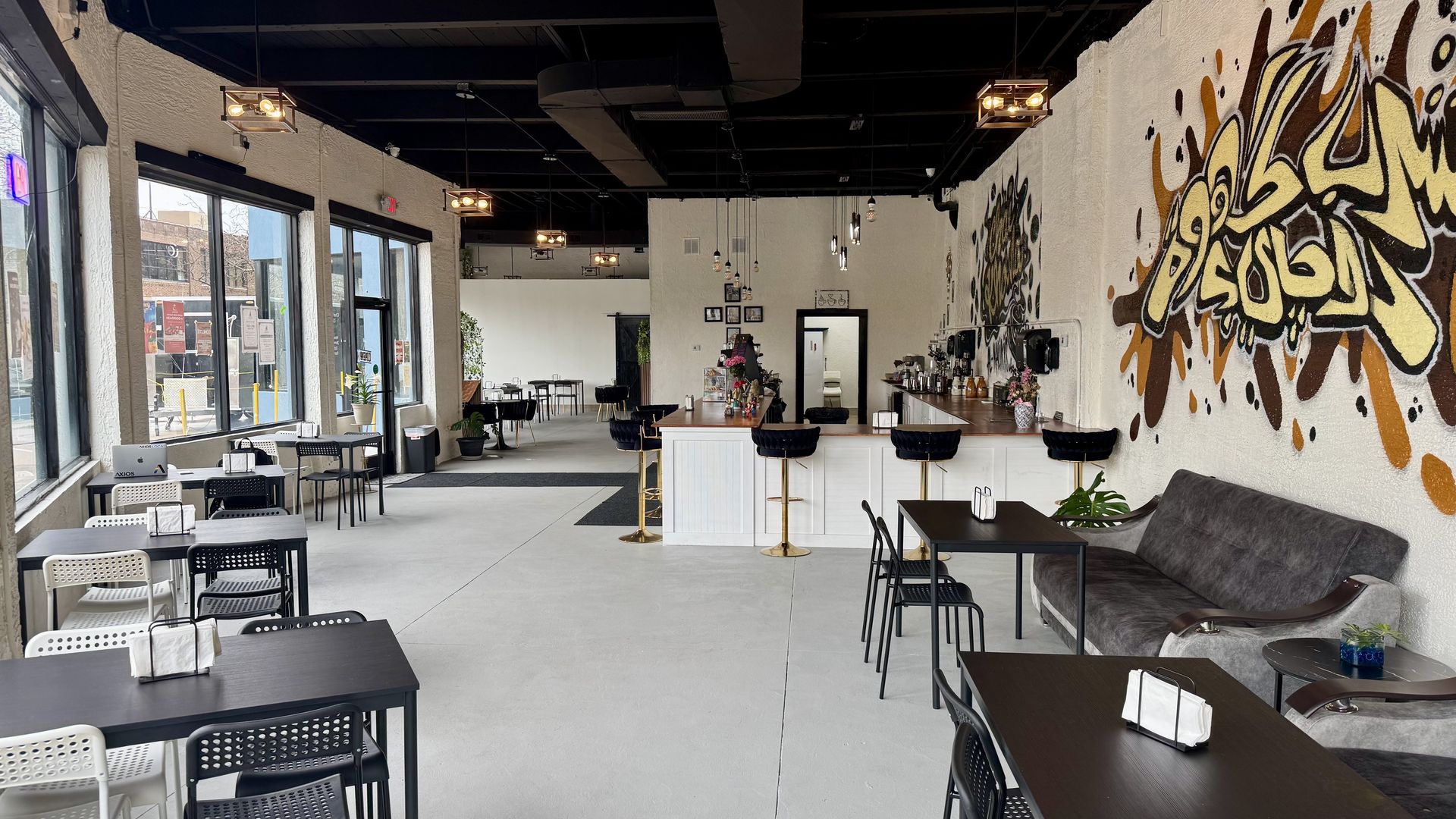 Bright cafe interior with a white bar and gold stools on the right, a bold yellow-brown graffiti mural on the wall, and rows of black and white tables by large windows on the left; exposed ceiling with pendant lights.