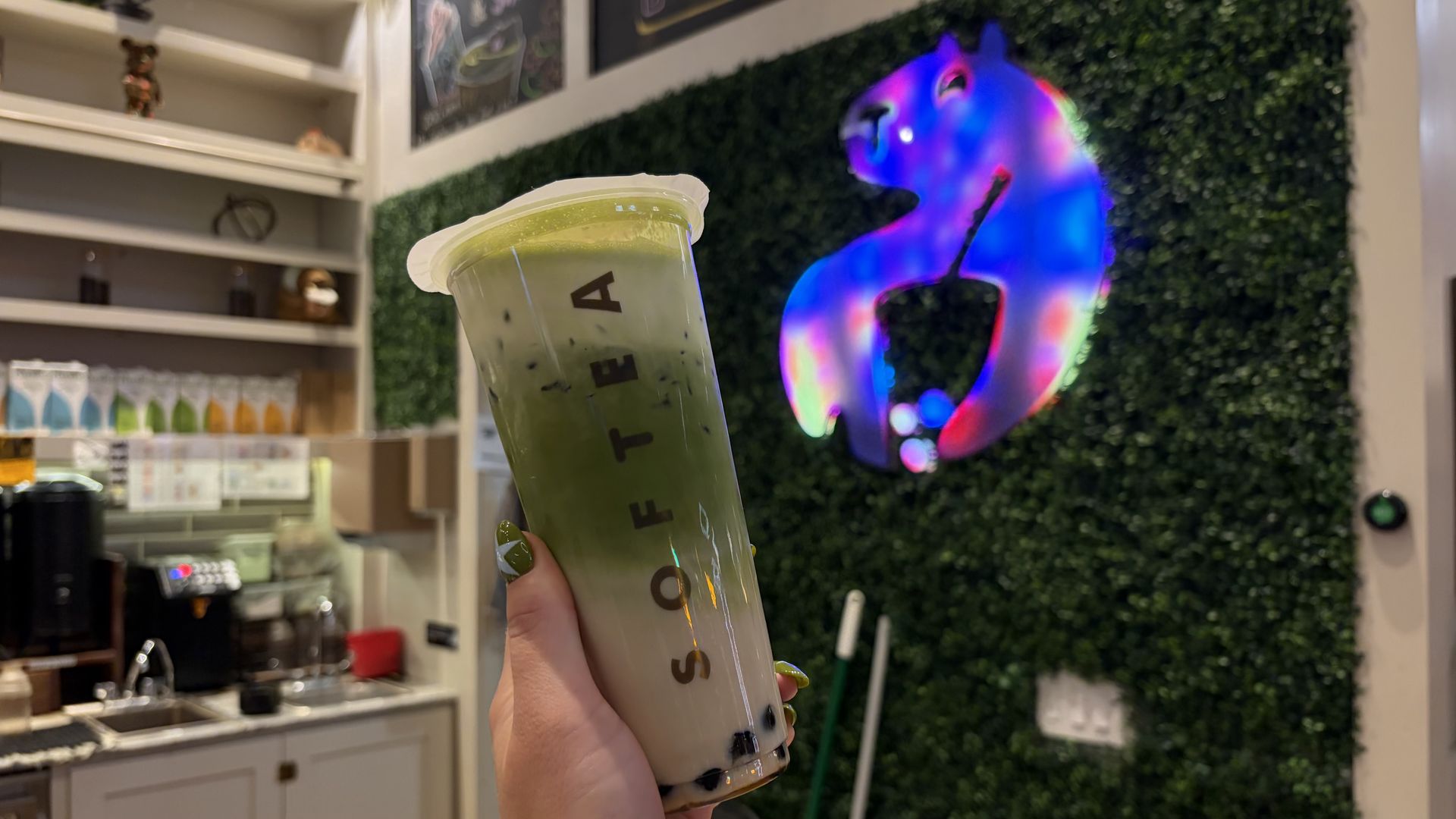 A hand holding a tall plastic cup of green iced matcha with a white lid, in a cafe with white shelves and a vivid neon unicorn mural on a green hedge wall.