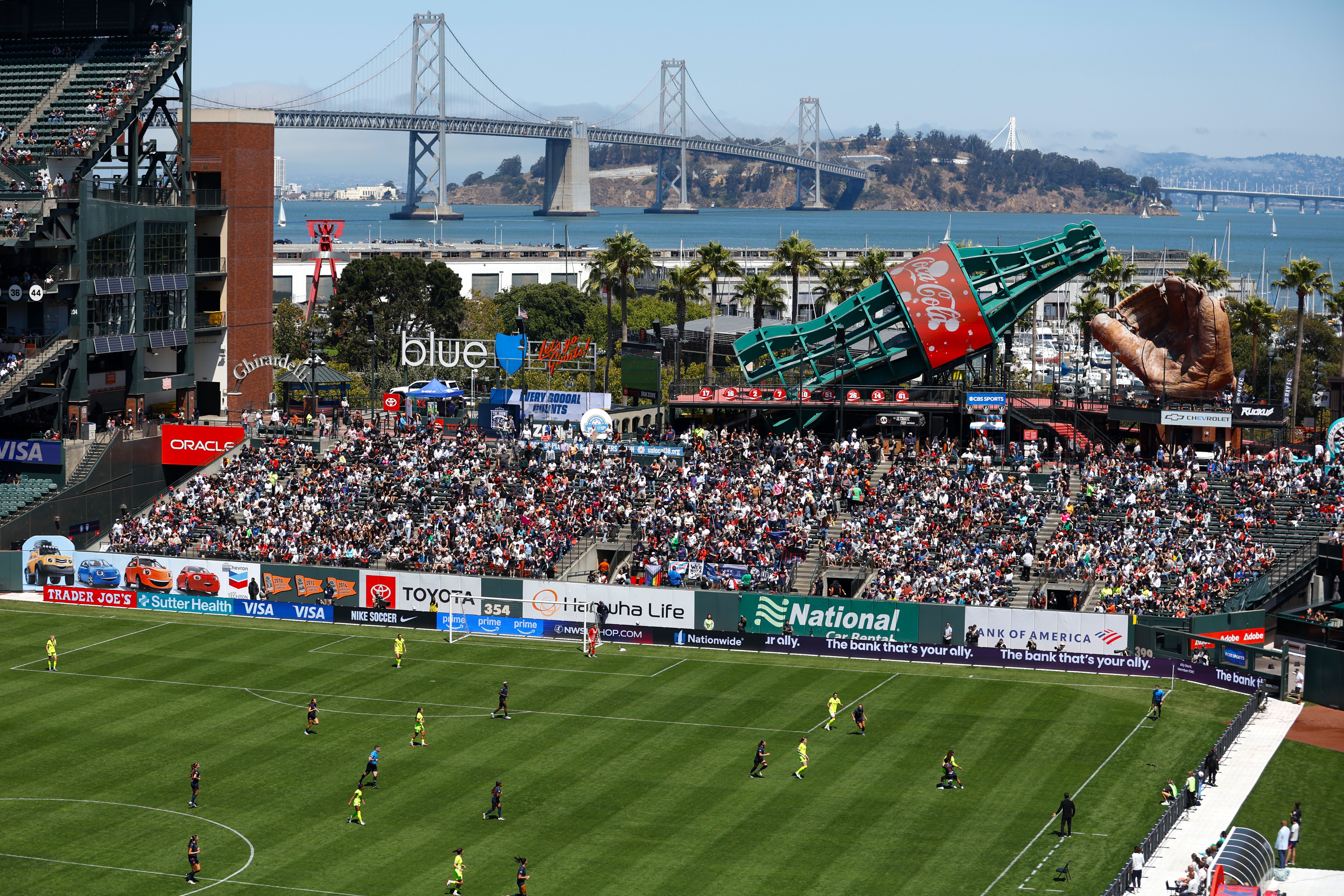 Soccer game with players on green field and crowd in stadium stands. Large Coca-Cola bottle and baseball glove sculptures. Bay Bridge and water in background under clear sky.