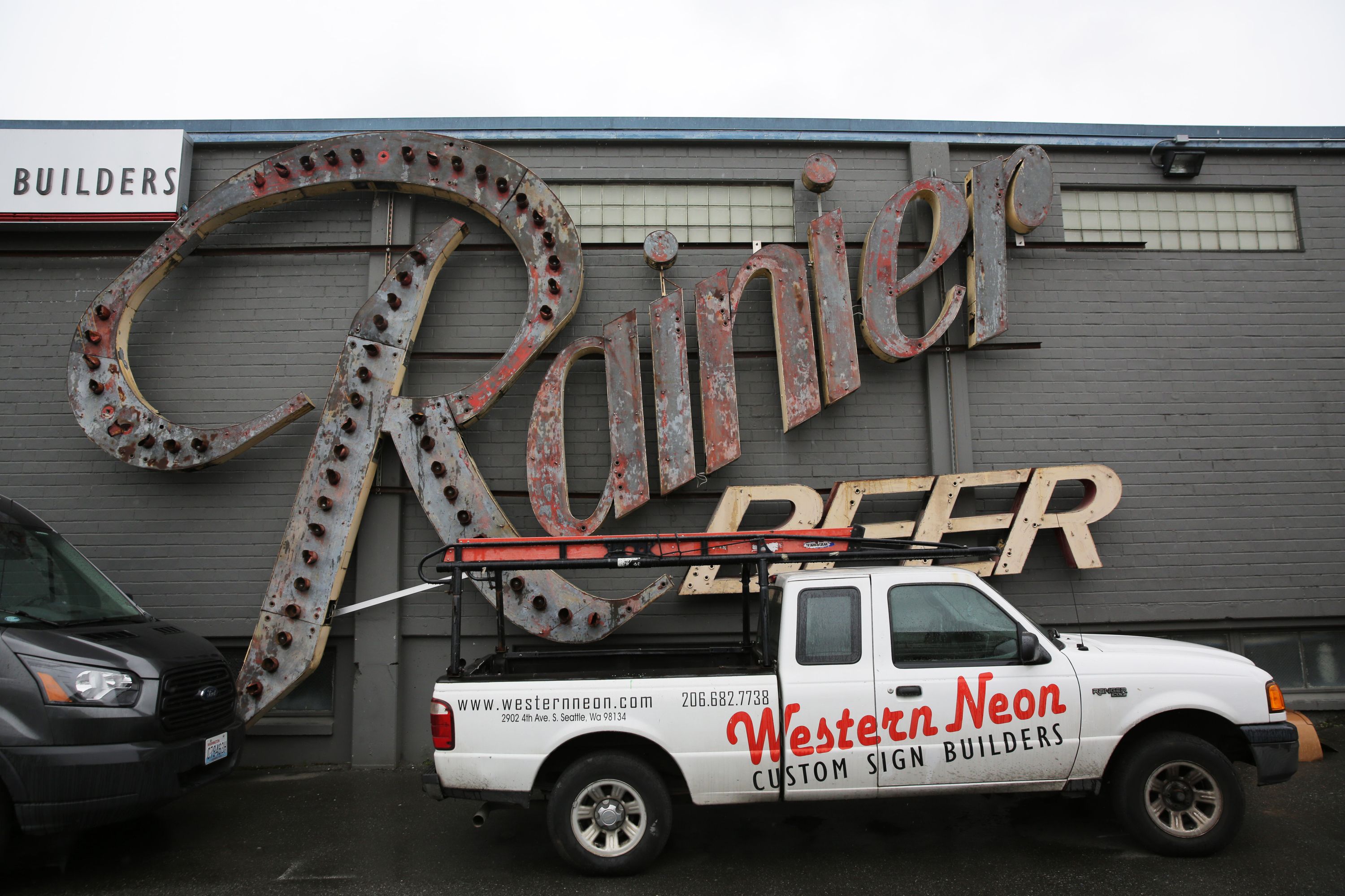 A fading Rainier Beer neon sign sits on a wall while a white truck with "Western Neon" written on the side is pulled up alongside it.