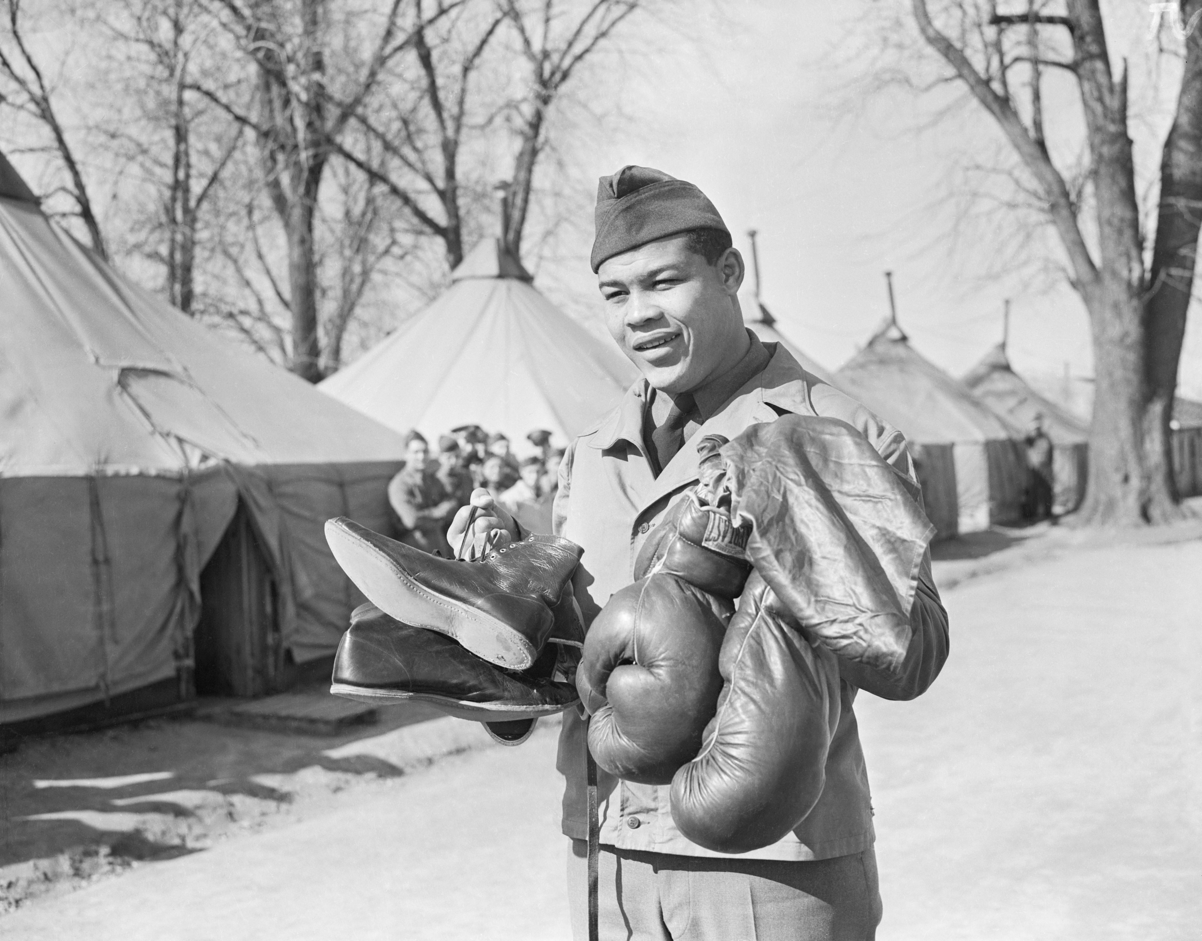 Joe Louis in uniform stands outdoors holding boxing gloves and boots, with military tents and leafless trees in the background on a sunny day.