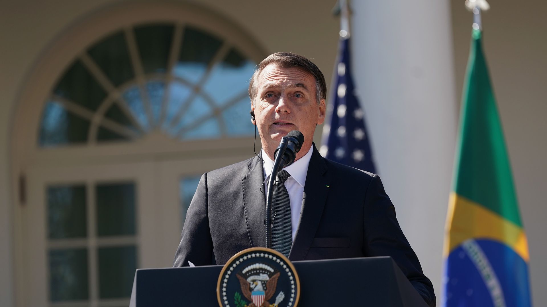 Brazilian President Jair Bolsonaro is pictured here speaking at a podium at the White House, with an American and Brazilian flag behind him.