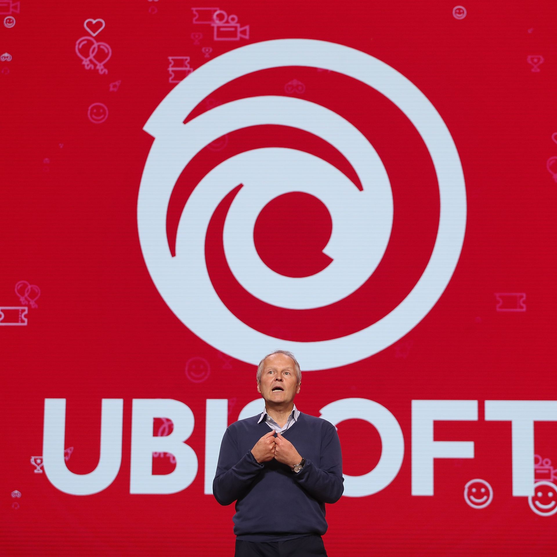 Photo of a man in pants, sweater, and a collard shirt standing in front of a big version of the Ubisoft logo