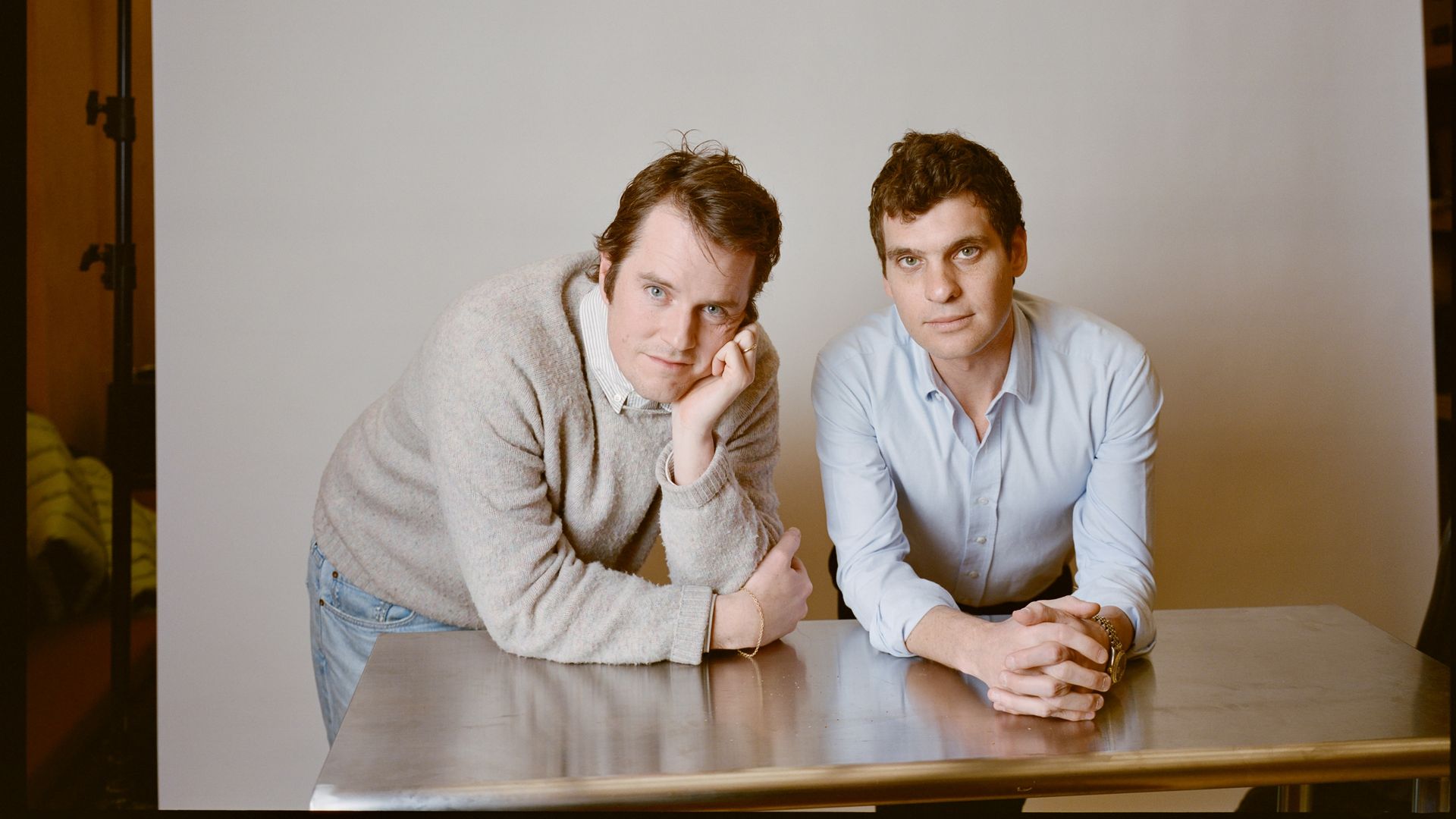 Two men lean on a metallic table against a plain light gray backdrop; one wears a beige sweater, light blue jeans, and the other a light blue shirt; both look directly at the camera.