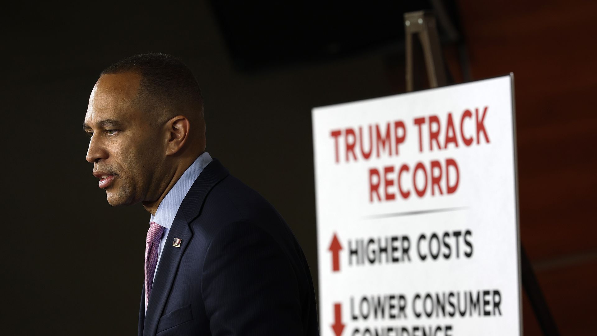 Hakeem Jeffries, wearing a blue suit and standing in front of a white poster with "TRUMP TRACK RECORD" on it.