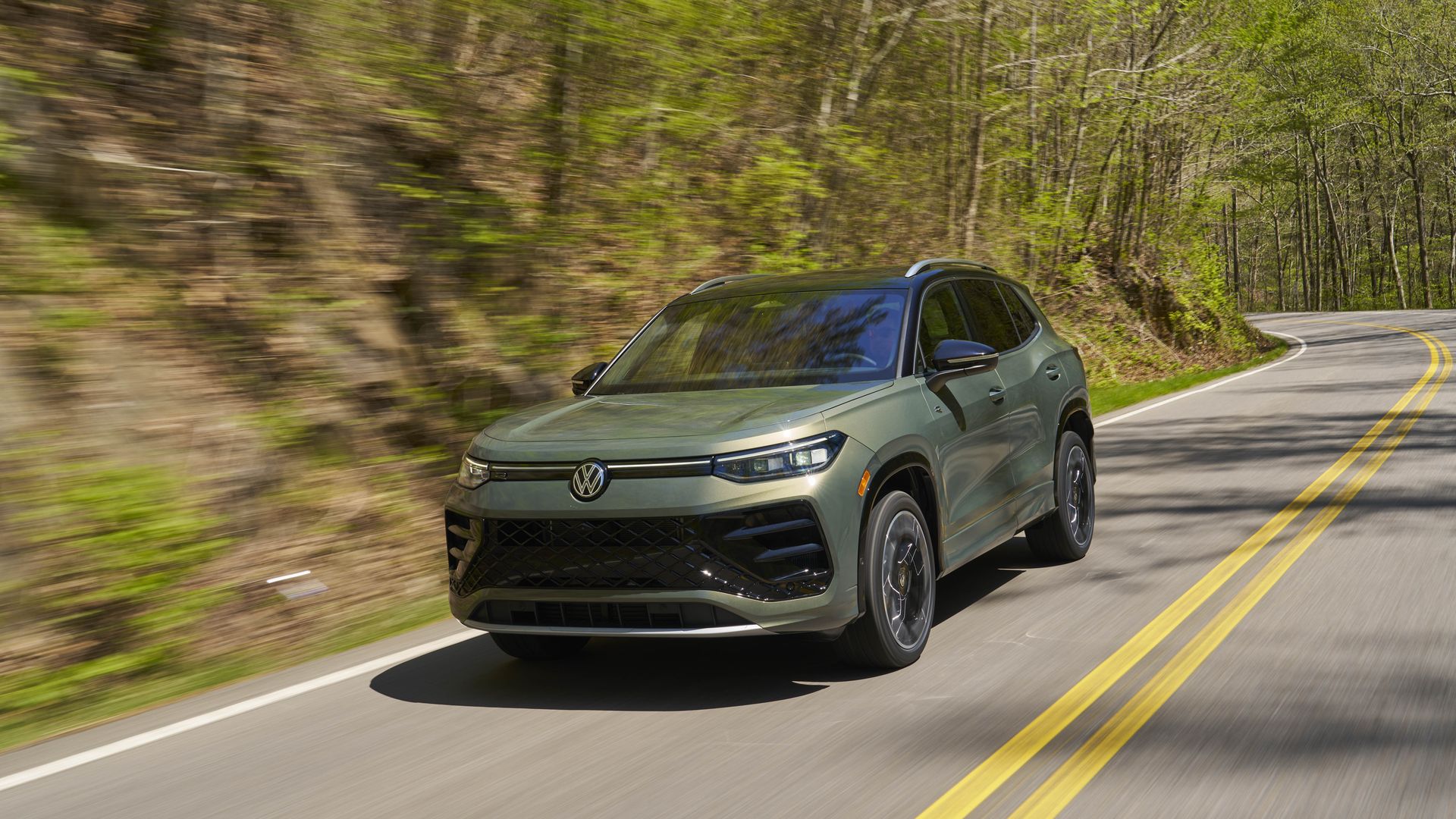 Green Volkswagen Tiguan SEL R-Line SUV speeds along a winding forest road, motion blur in the trees, with a double-yellow center line and curved pavement under bright daylight.