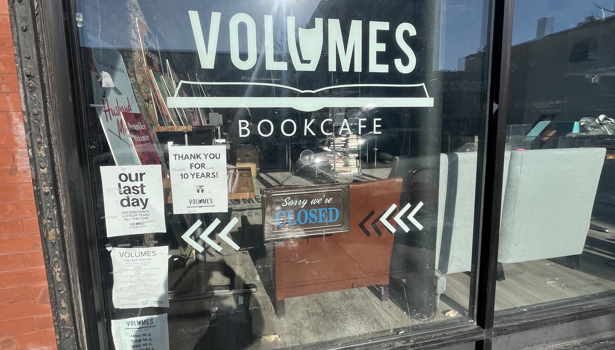 Front window of Volumes Bookcafe with signs stating "our last day", "Thank you for 10 years!", and a hanging sign "Sorry we're CLOSED". Inside chairs and stacks of books visible.