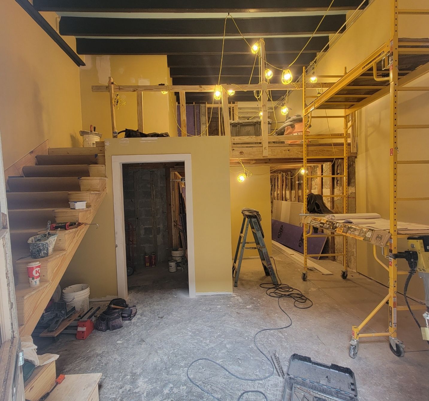 The back of Manolito is seen under construction. A stairway leads to a lofted seating area above, and wooden beams are visible in the ceiling. Scaffolding and temporary hand railings are visible.