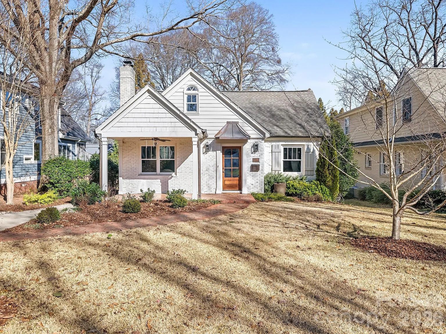 White brick single-story house with wooden front door, covered porch, and leafless trees in front yard, under a clear blue sky on a sunny day.