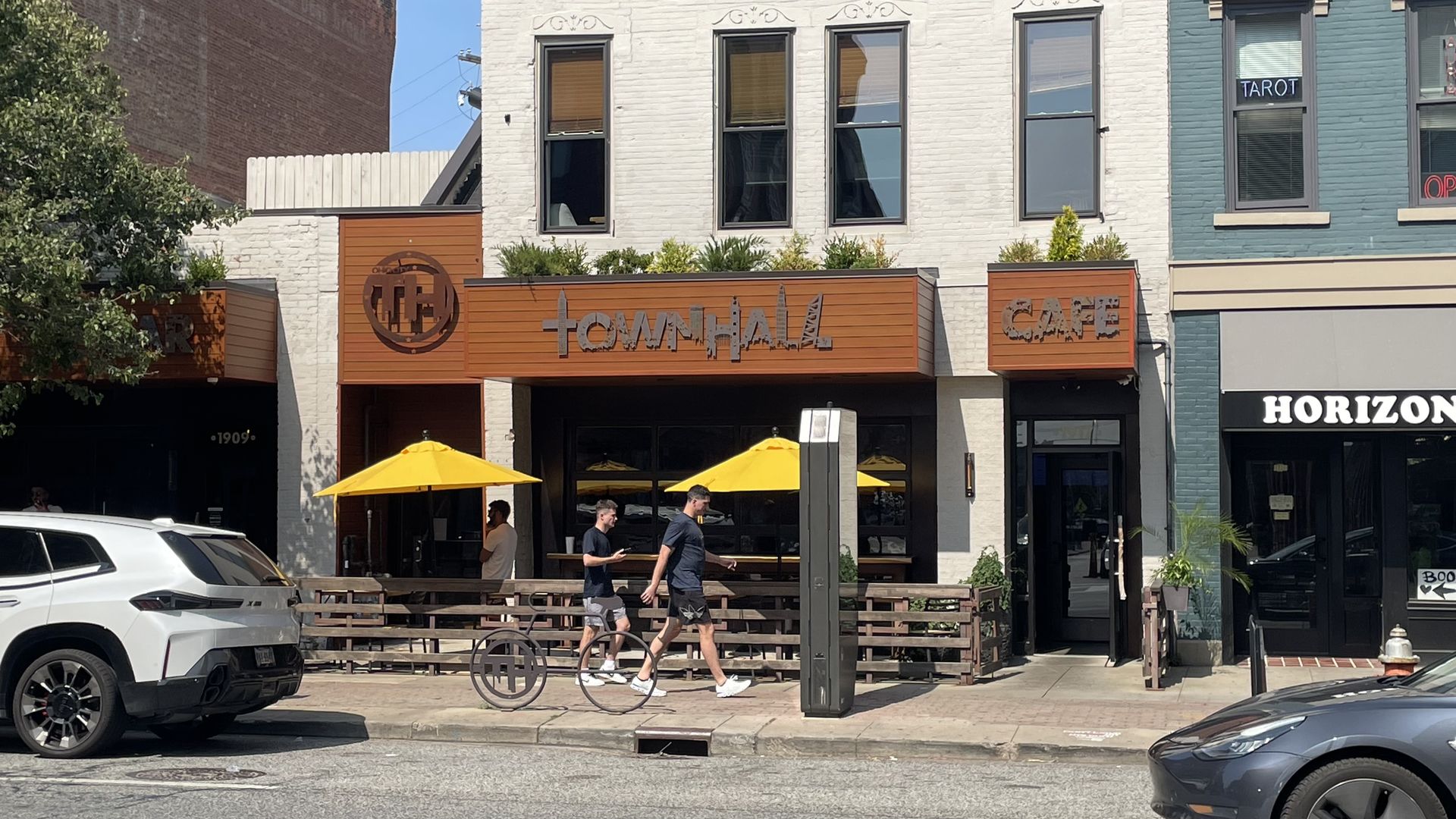 Sunny street view of a white brick building with orange storefront reading "Town Hall Cafe"; yellow outdoor umbrellas shade tables, pedestrians walk by, and cars are parked along the curb.