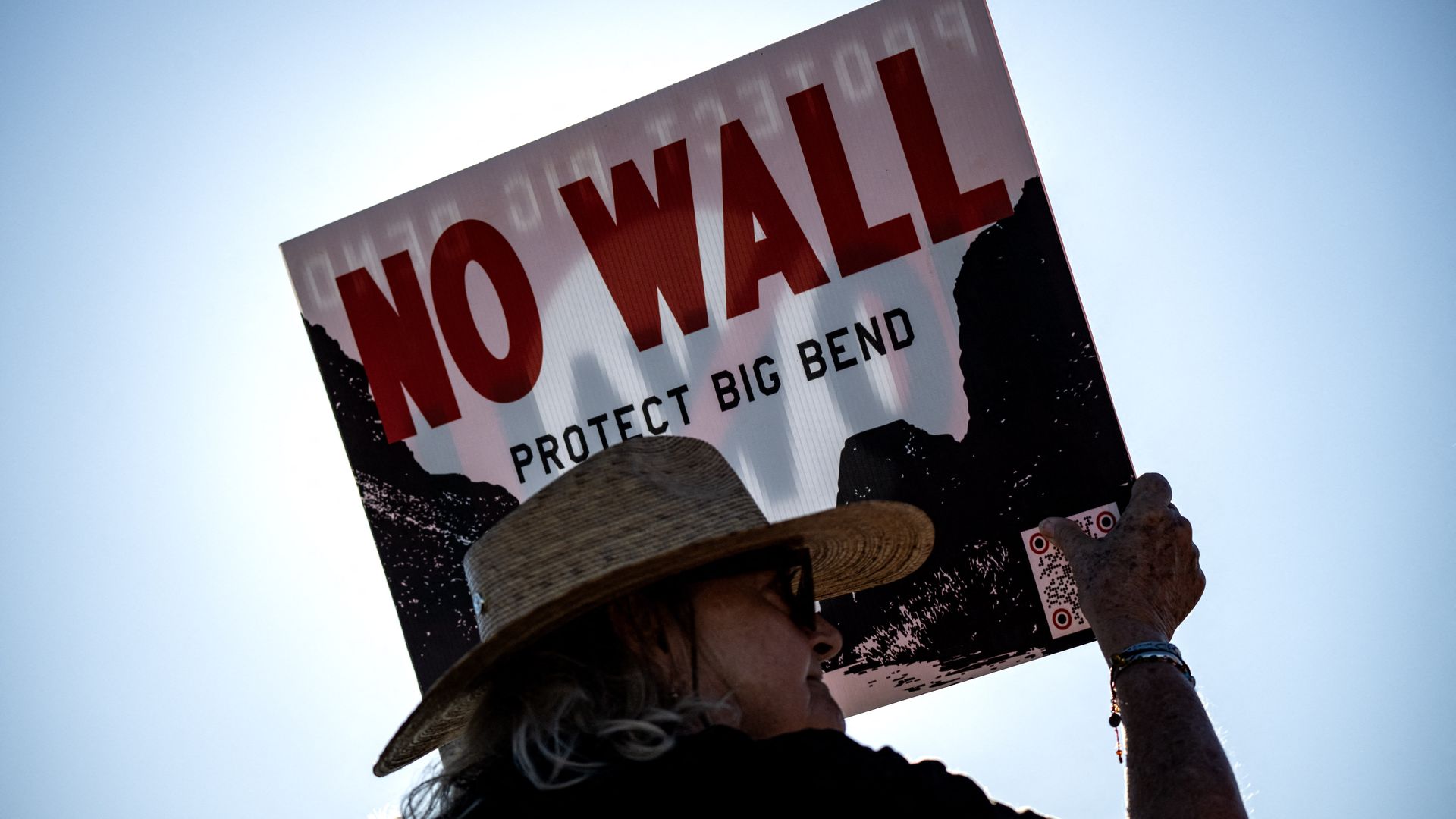 A woman wears a straw hat and glasses while holding a sign that reads "NO WALL PROTECT BIG BEND."