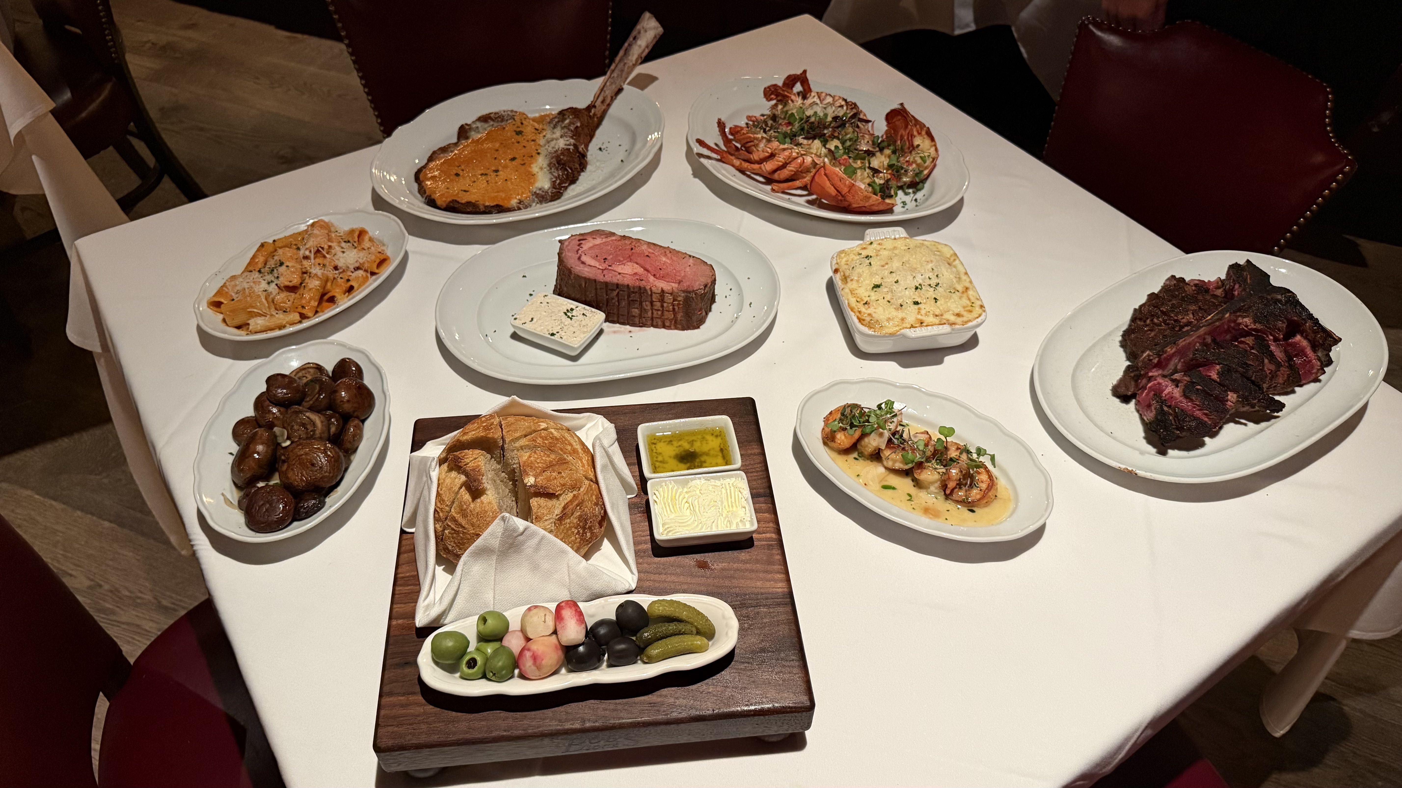 White table with assorted dishes including sliced rare roast beef, grilled lobster halves, roasted mushrooms, shrimp in sauce, pasta with cheese, bread with olives, pickles, butter, and dipping oil.