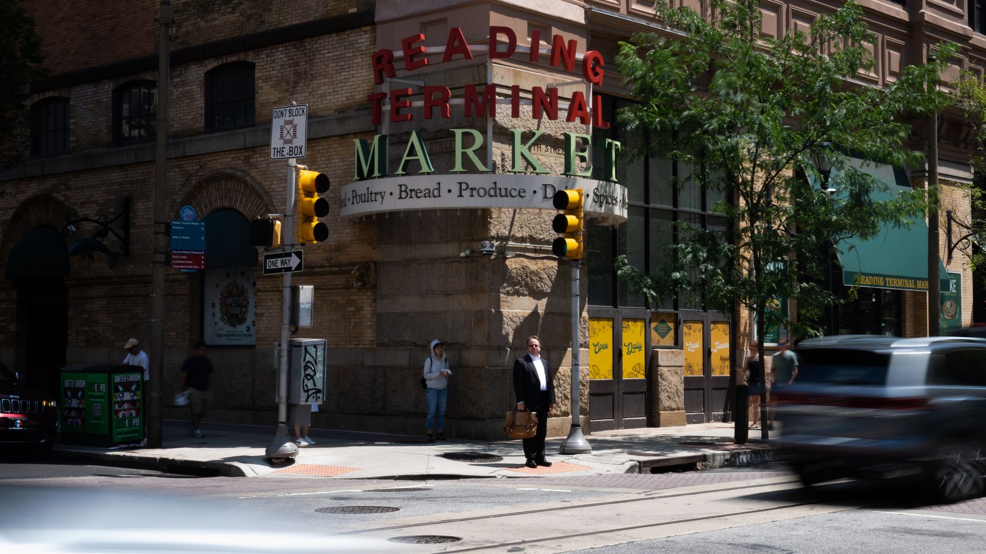 Street corner outside Reading Terminal Market with people waiting to cross, brick building, traffic lights, and blurred cars passing by on a sunny day.