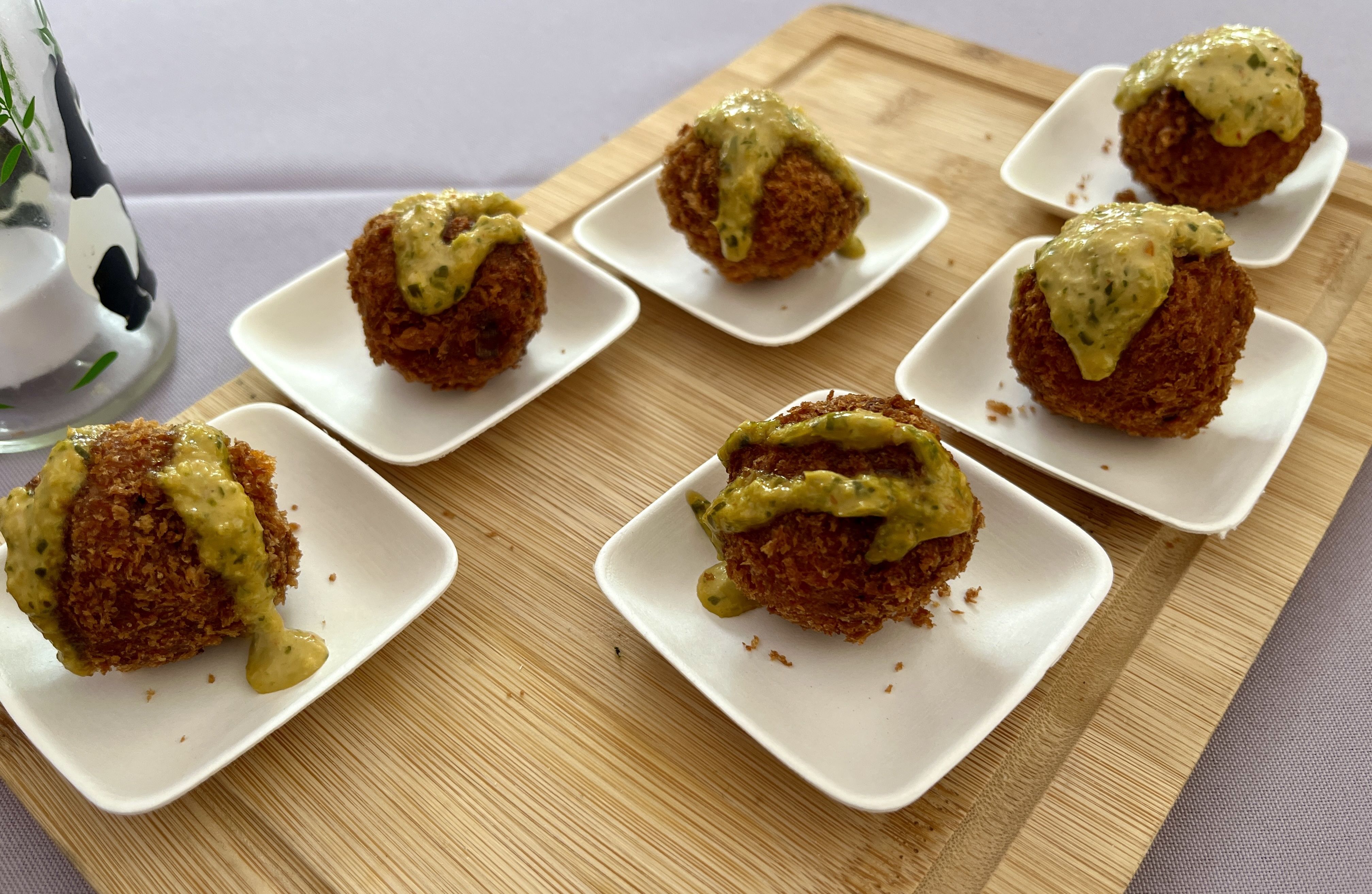 Golden fried croquettes topped with pale yellow herb sauce on small white plates arranged on a wooden board; a decorative vase with greenery is visible on the left.