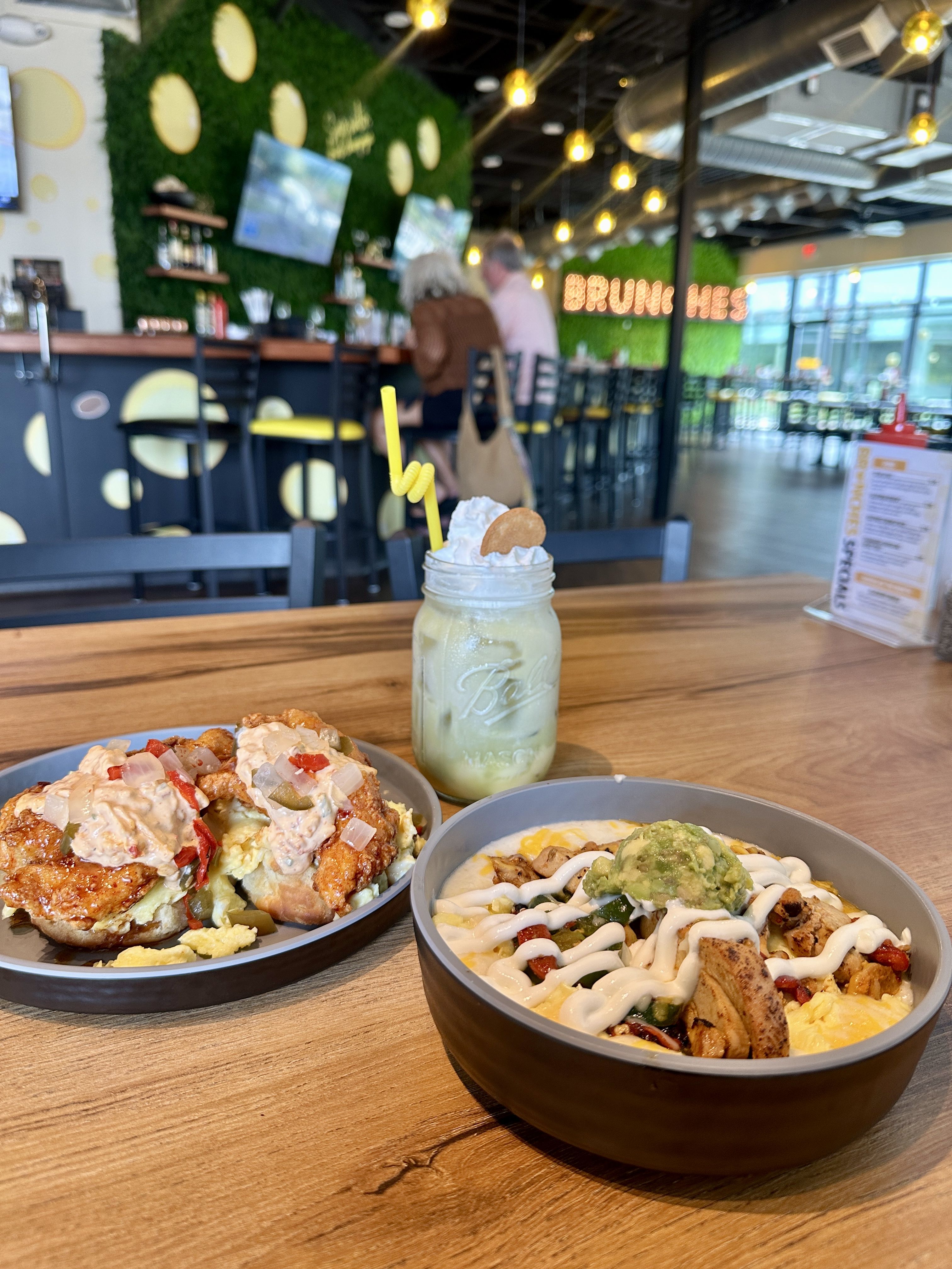 Brunches. Modern restaurant interior with a green wall and yellow circles. Foreground shows loaded nachos, a burrito bowl, and a mason jar drink with whipped cream and a yellow straw; blurred bar in back.