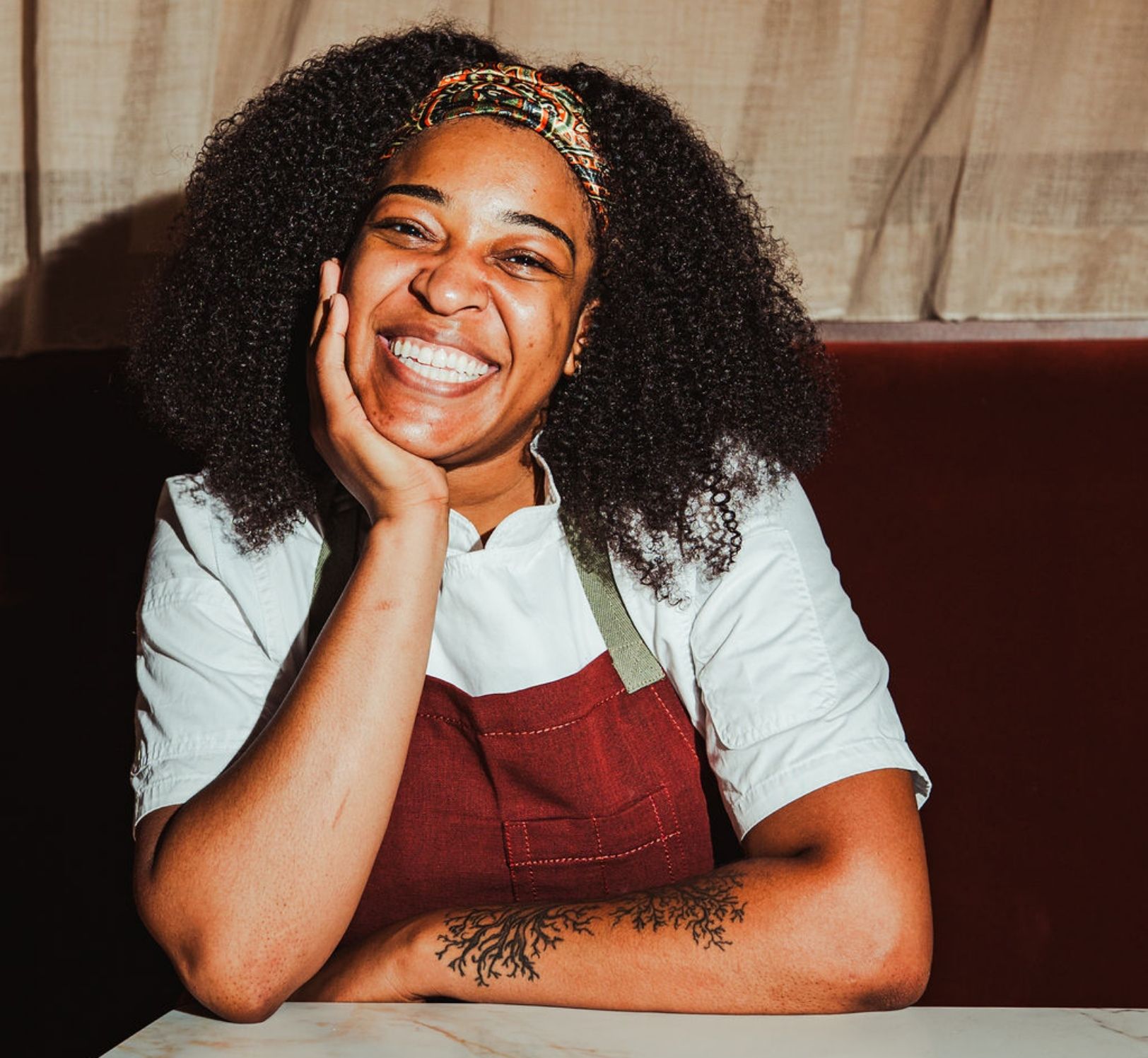 Smiling woman with curly hair and a patterned headband, wearing a red apron over a white shirt. She rests her cheek on her hand, with a tattooed forearm visible against a neutral background.