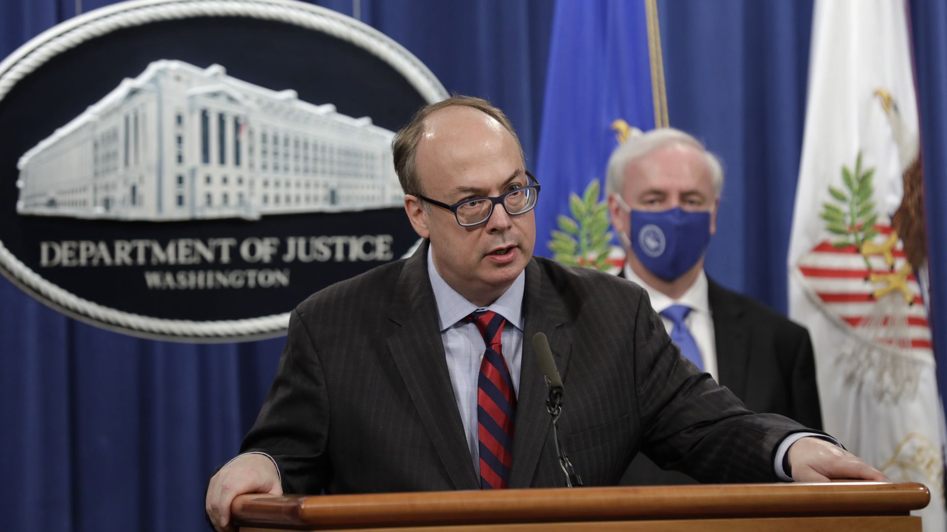 Man in suit and glasses stands at podium/lectern in front of a blue backdrop with The White House emblem.