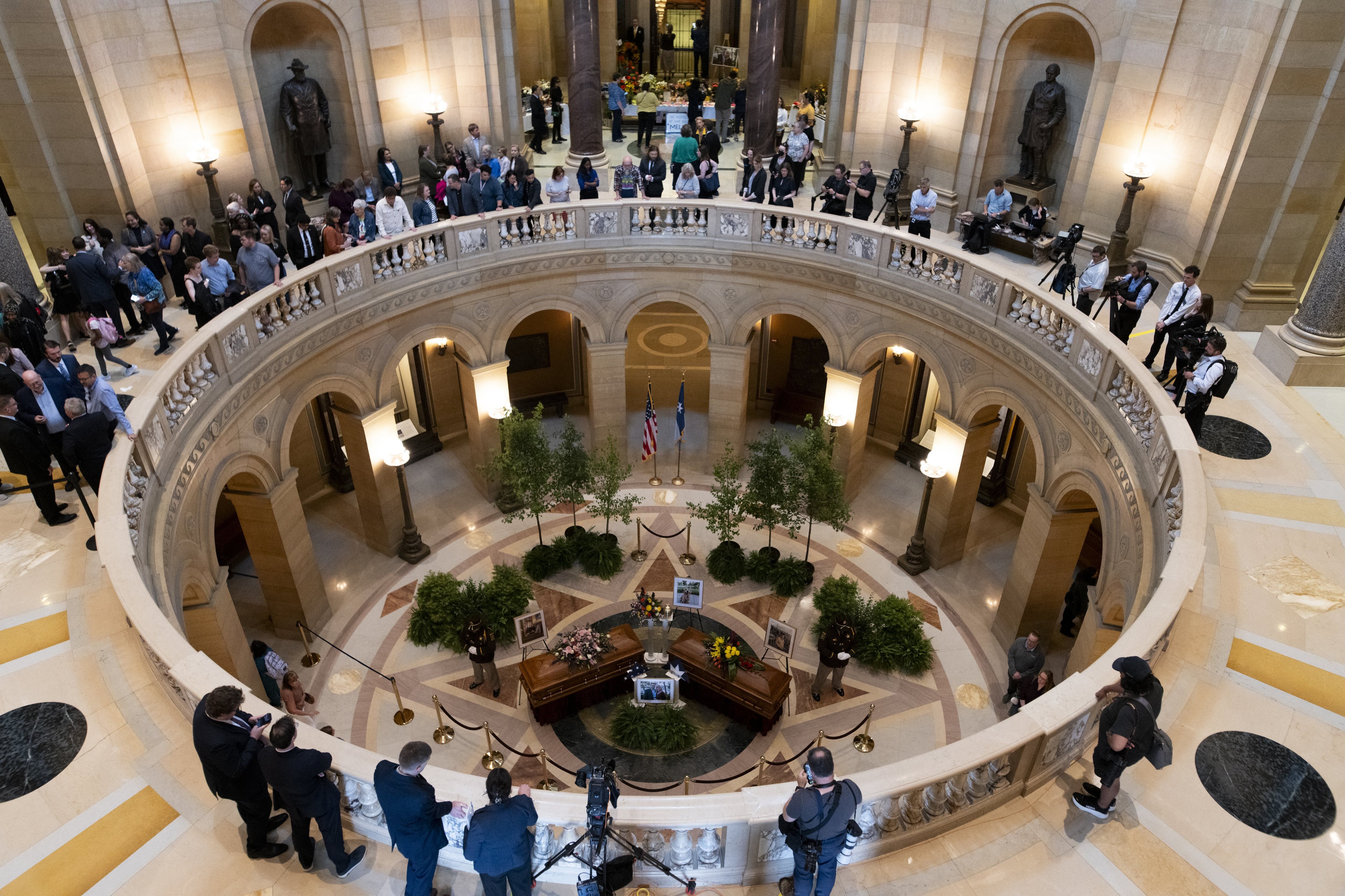 Interior of a grand circular marble atrium with a crowd along the curved balcony. Reporters with cameras stand to the right; a funeral display with flowers and a coffin on the lower level, flags nearby.