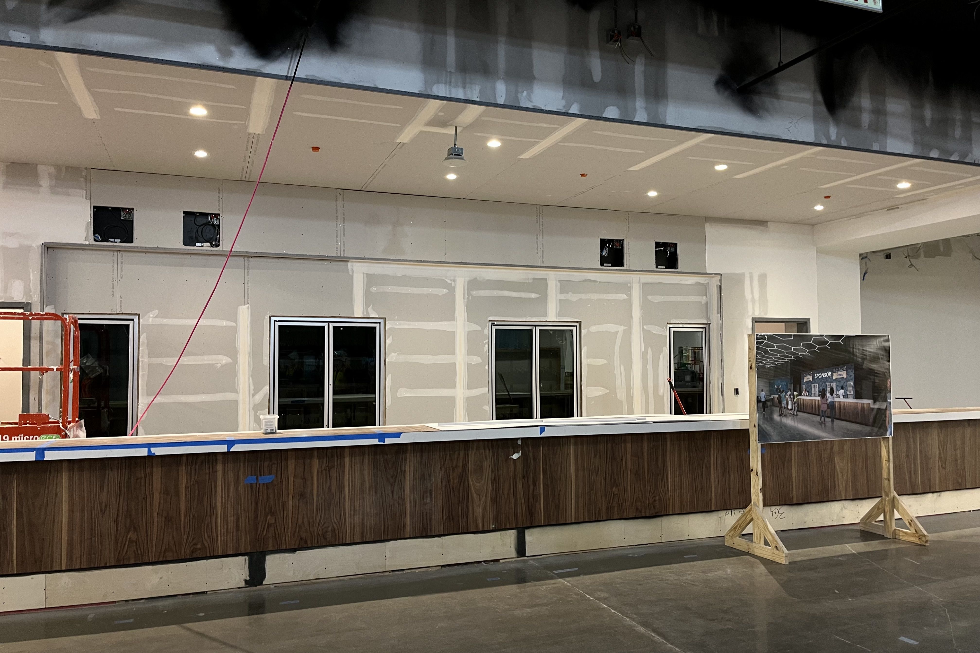 Interior of a construction site with a long wooden counter, unfinished white walls, recessed lighting, and a display board showing a future design with people at a sponsor booth.