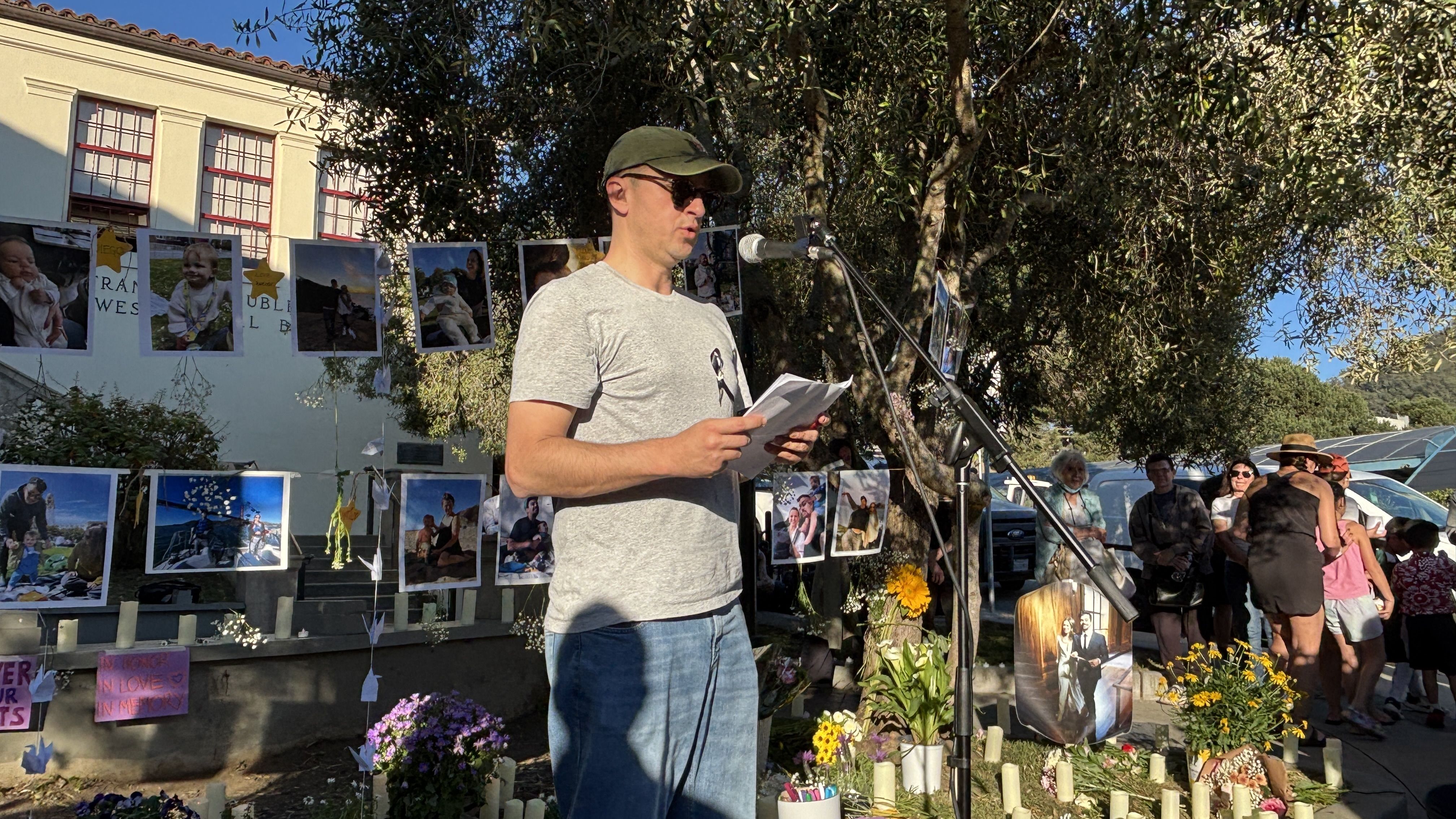 A man in a gray T-shirt, green cap and sunglasses speaks into a microphone, holding papers at an outdoor memorial with photos hung on a line behind him and a small crowd nearby.
