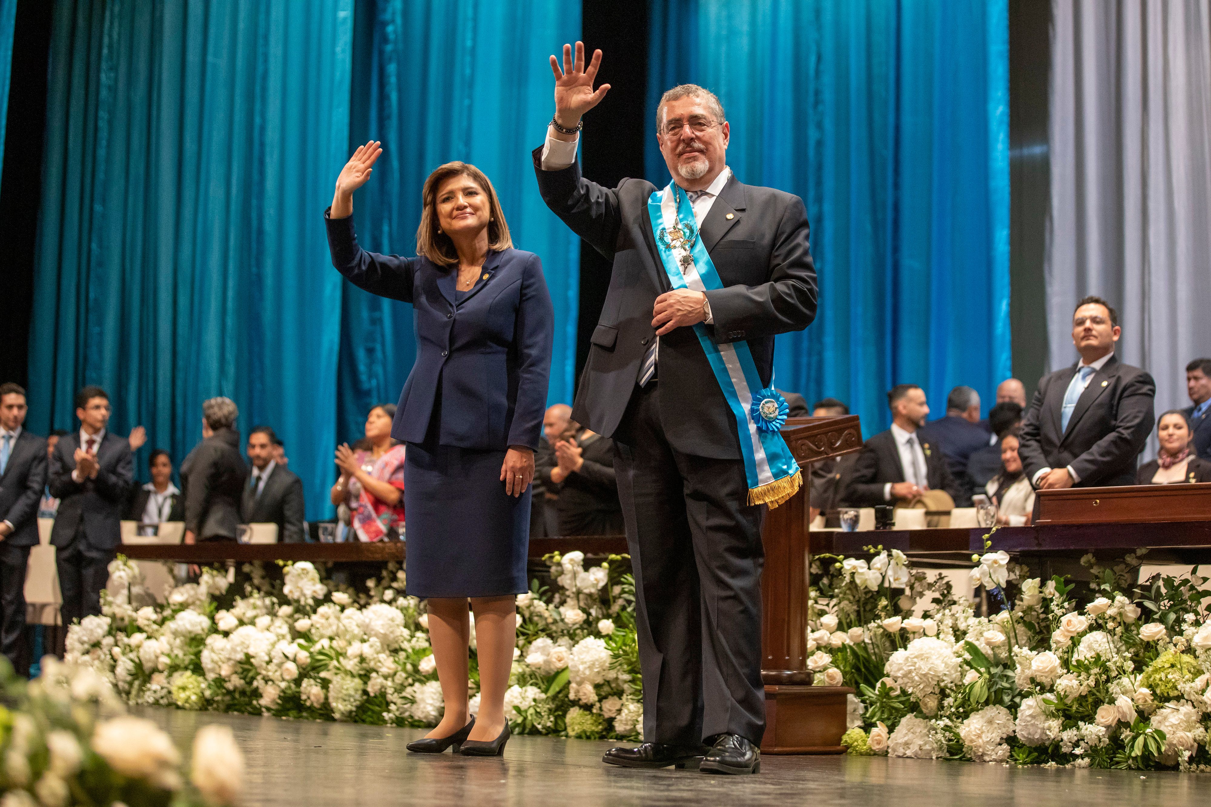 Guatemala president Bernardo Arévalo, right, and Vice President Karin Herrera wave with their left hands in front of a blue background with white flowers during their inauguration.