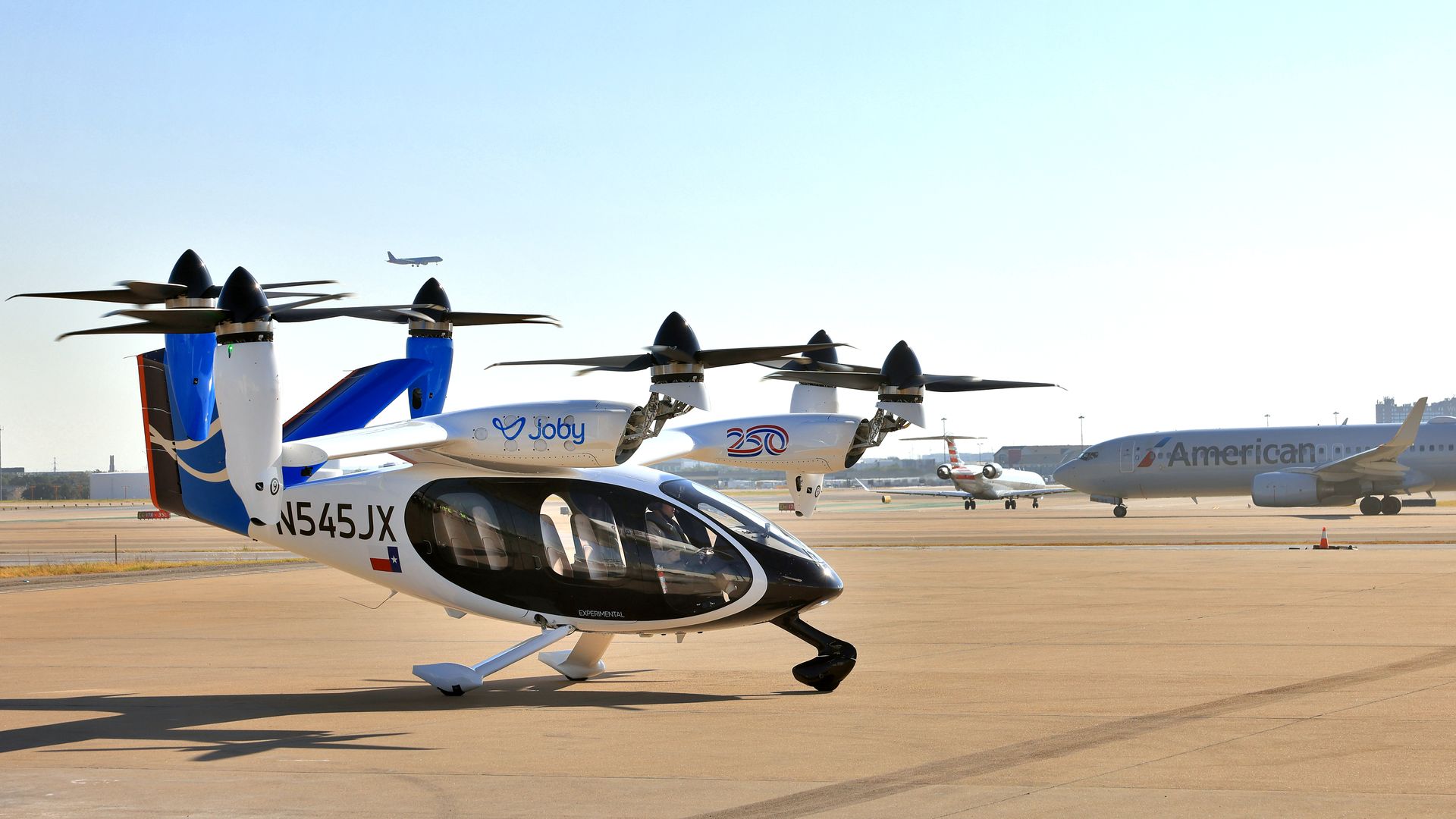 An electric air taxi sits on a runway with an American Airlines plane behind it