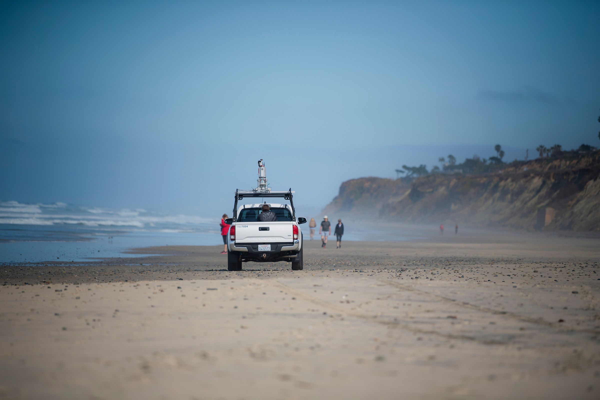White pickup truck with equipment on beach near ocean waves, people walking and standing on sand, cliffs and blue sky in background, mist along coastline.