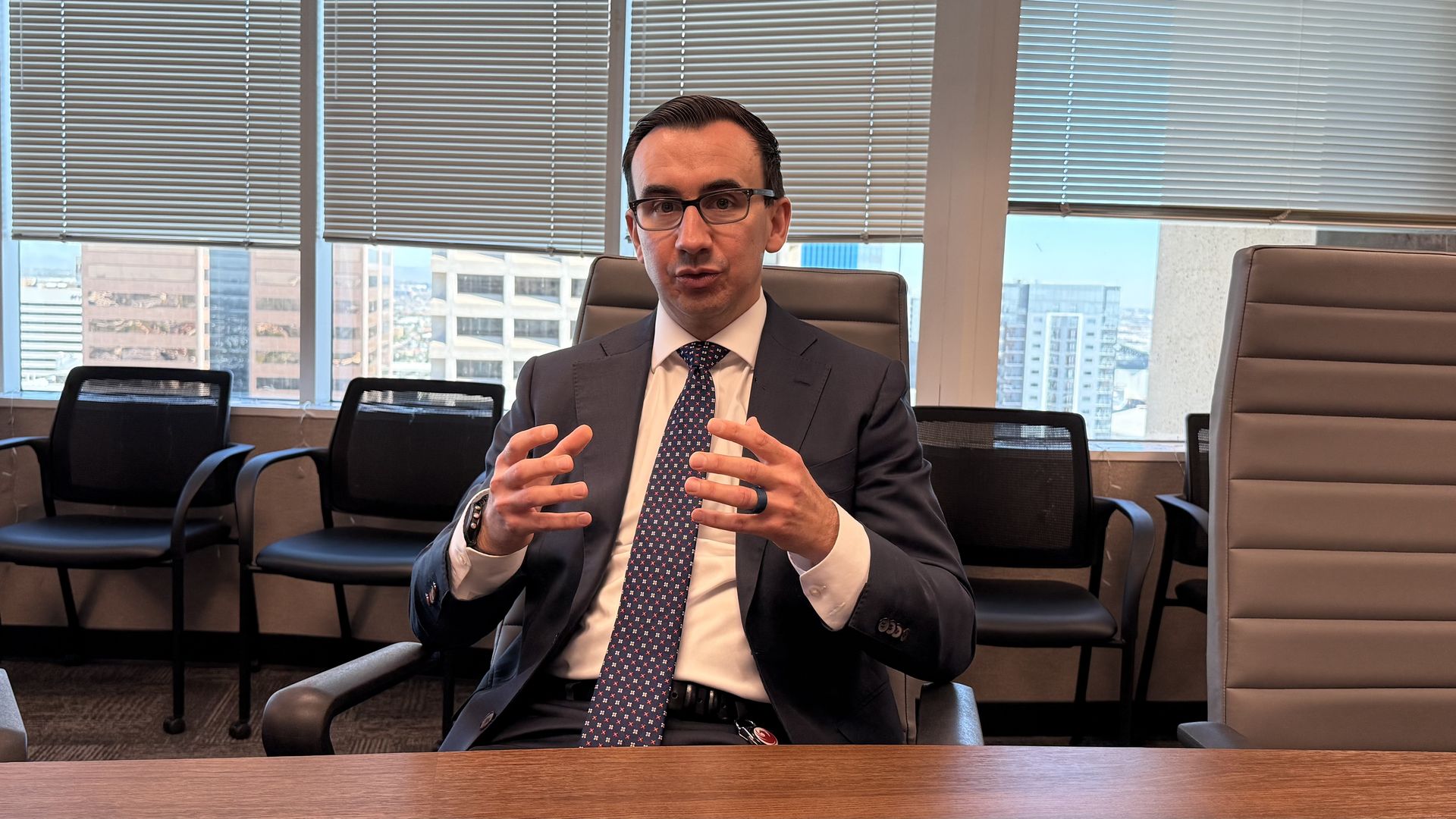 Man in dark suit, white shirt, and patterned tie speaking and gesturing with hands, sitting in a chair in a conference room, with high-rise buildings visible through the large windows. 