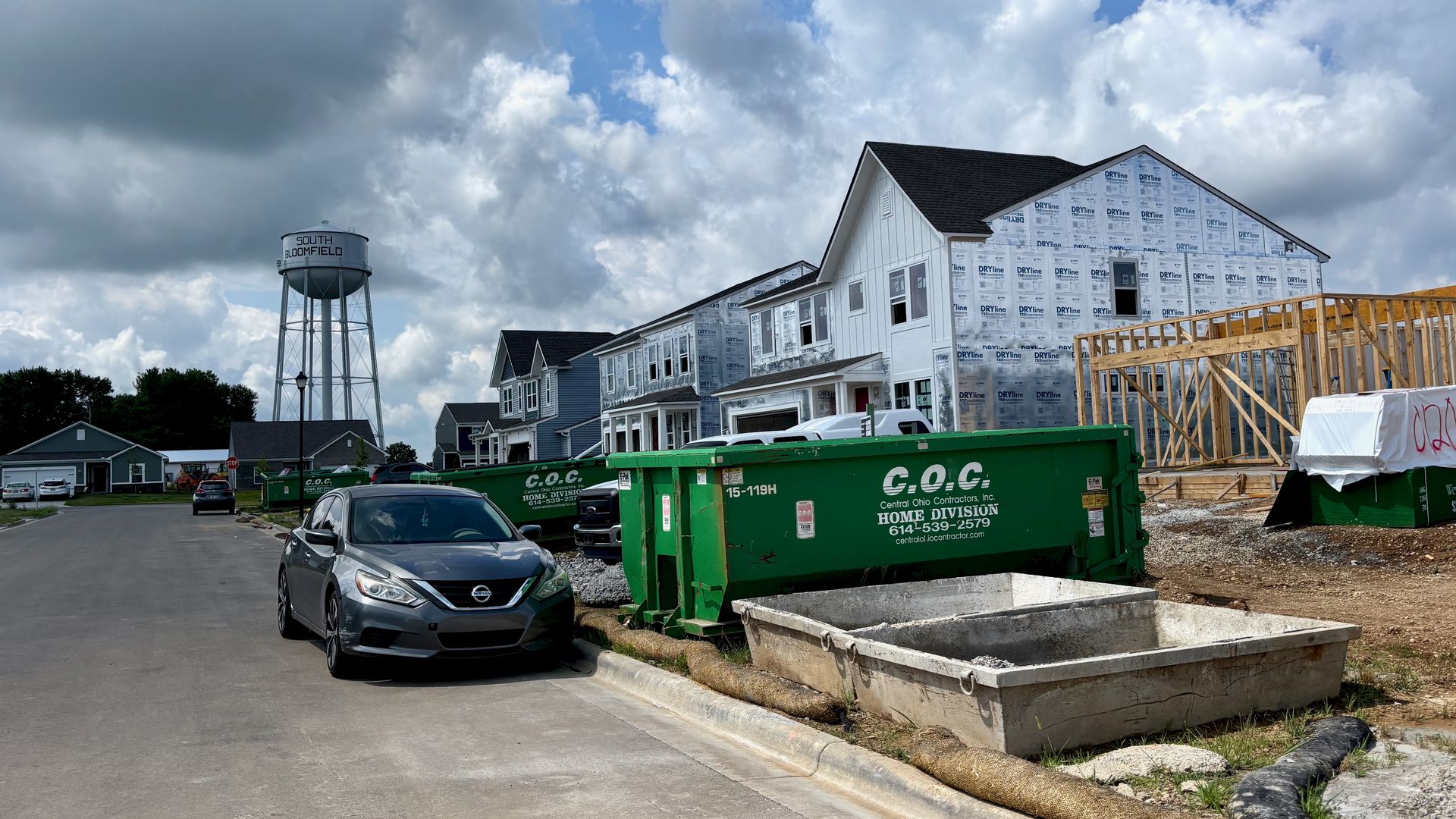 Suburban construction site: a two-story house wrapped in white sheathing with visible framing, a green C.O.C. dumpster, and a grey car on the street, under a cloudy blue sky and a distant water tower.