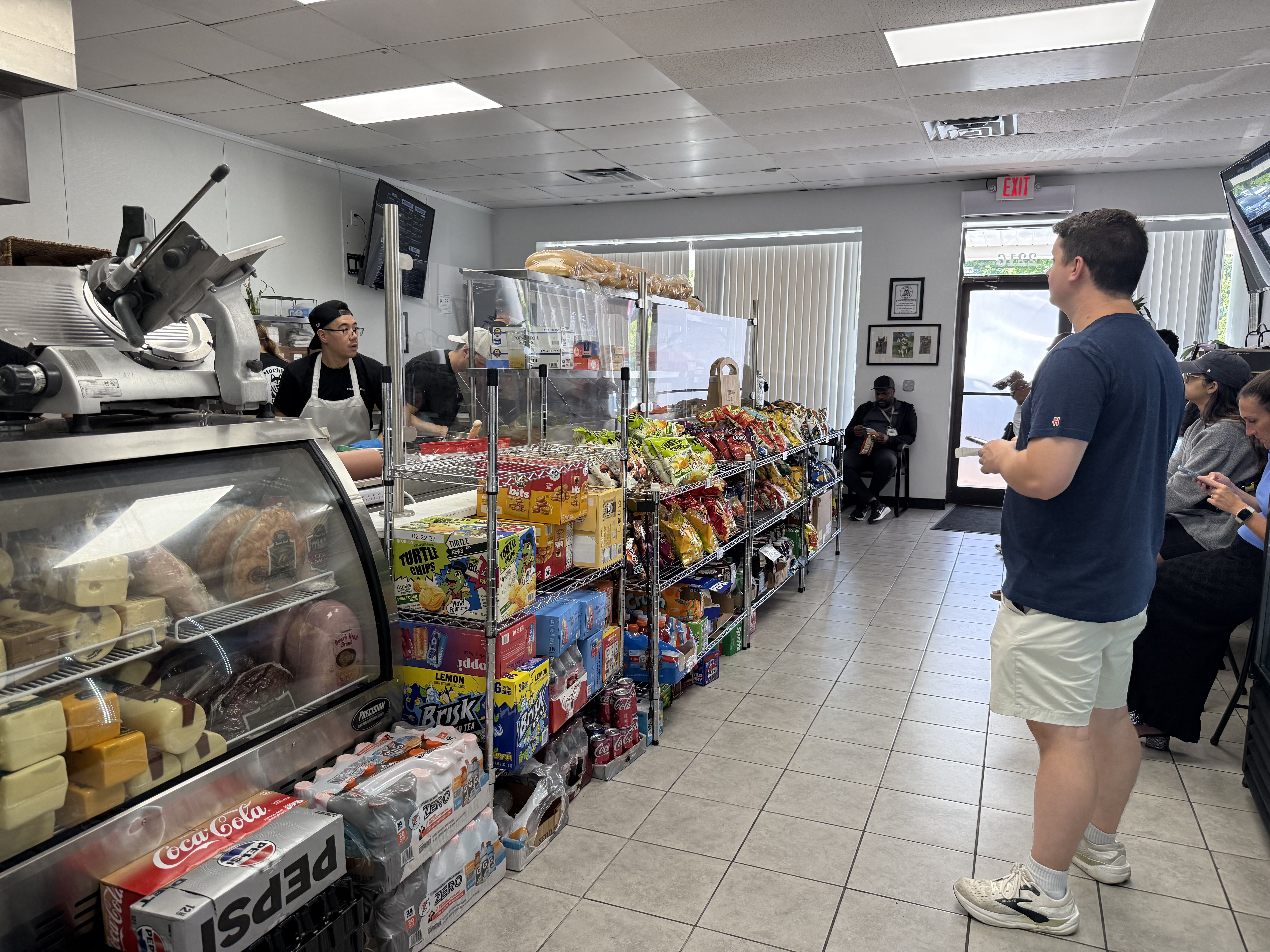 Inside a casual deli, a large meat slicer sits at the left counter. A glass cheese display and snack shelves line the middle as customers wait; a man in a blue shirt faces away near the exit.