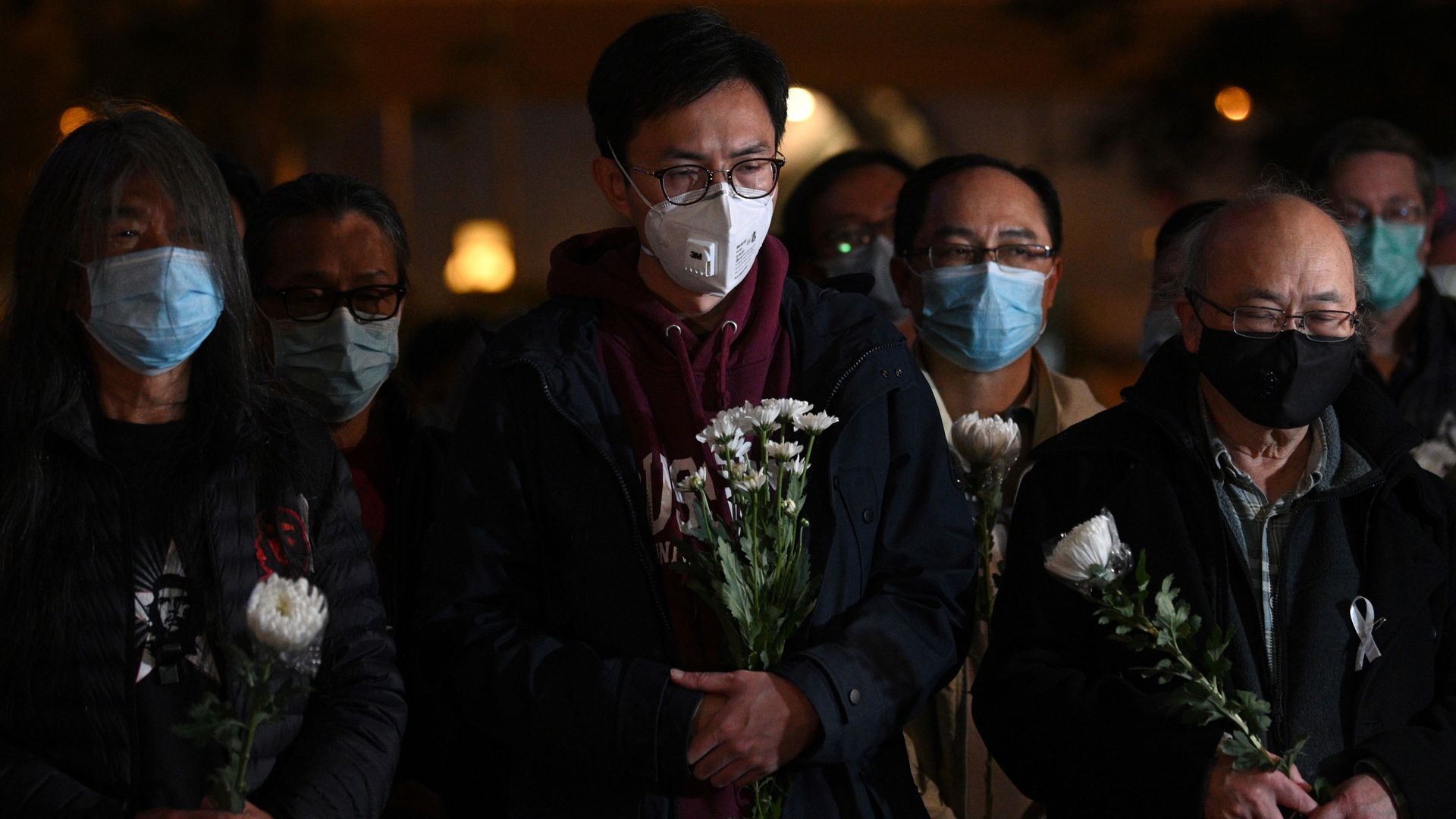 In this image, several people in Hong Kong wearing face masks hold flowers and stand in a silent vigil outside at night