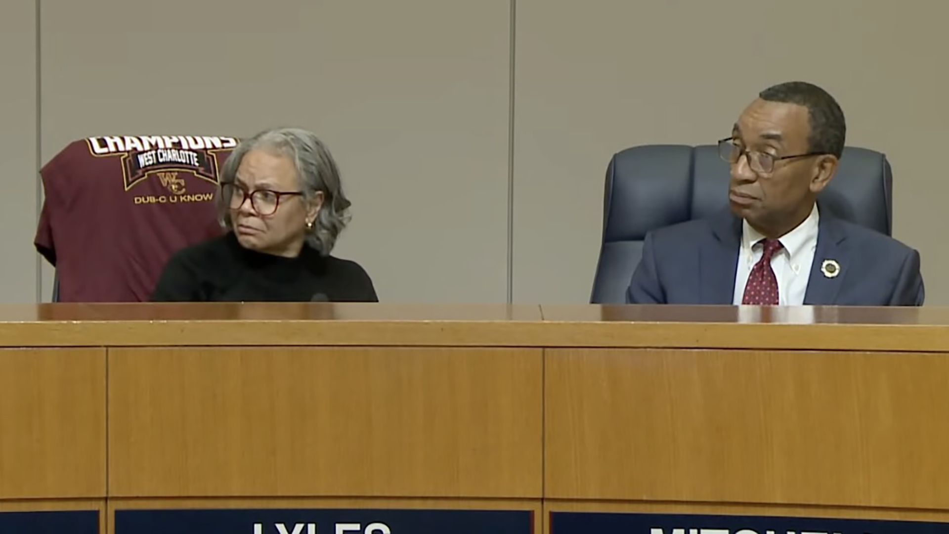 Two people sit at a wooden panel desk: a gray-haired woman with red glasses on the left and a man in a suit and red tie on the right; a maroon chairback behind reads "Champions West Charlotte".