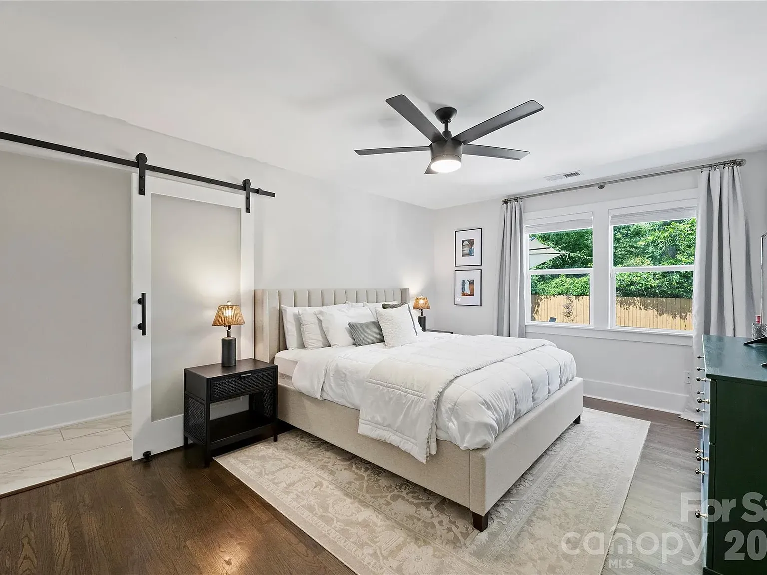 Bright modern bedroom with a beige upholstered bed and white comforter, two black nightstands with lamps, sliding barn door on the left, a ceiling fan, and a large window with gray curtains, plus a green dresser.