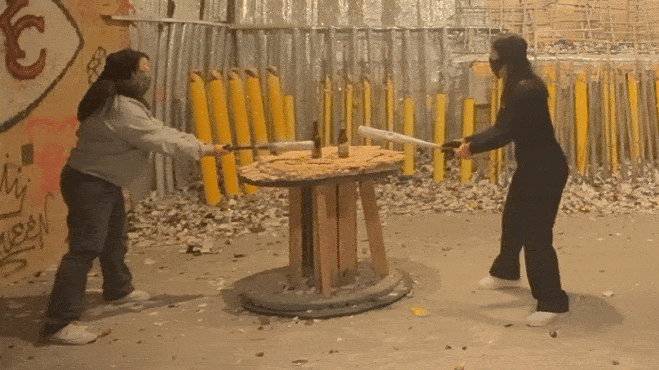 Two people wearing masks and holding bats stand on opposite sides of a wooden table with two bottles in an industrial setting with yellow poles and debris on the ground.