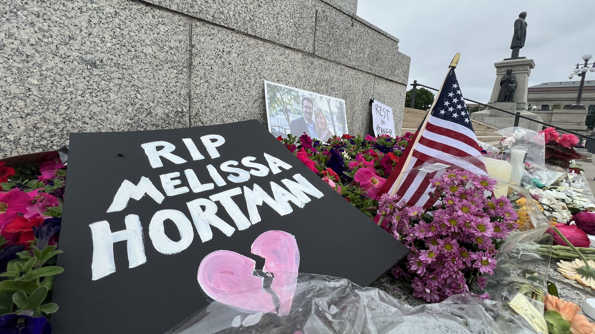 Memorials to a fallen state lawmaker line the base of a stone monument. Visible are flowers, an American flag, a portrait of a couple and a sign reading "RIP Melissa Hortman."