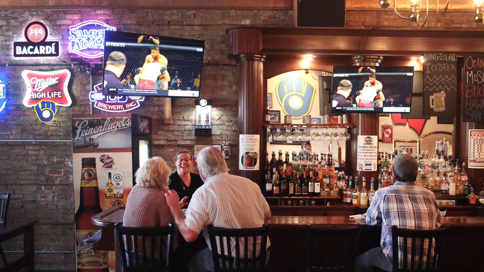 In this image, a couple sits next to each other at a bar with one chair between them and a man sitting nearby